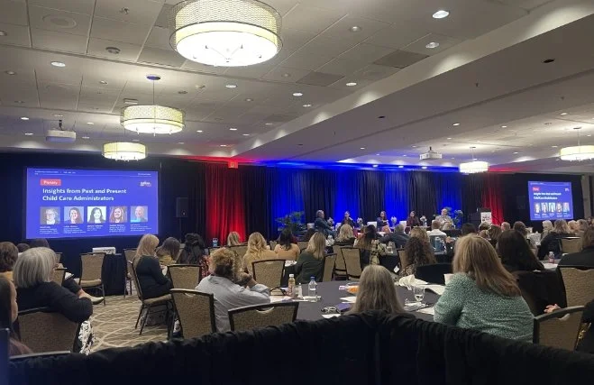 Conference room filled with attendees watching a presentation on child care insights, with large screens and a stage decorated with red, white, and blue curtains.