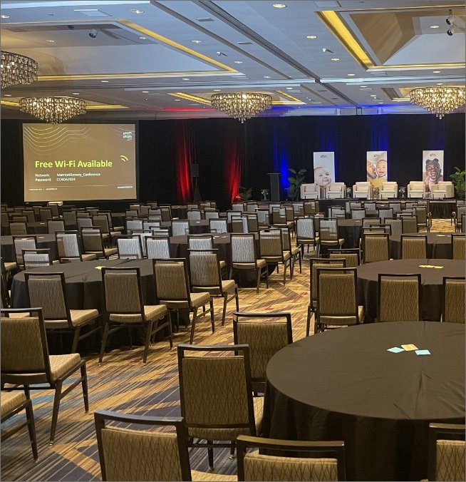 Empty conference room with round tables and chairs, a stage with curtains, large screen displaying Wi-Fi information, and portraits on stands, with bright chandeliers overhead.