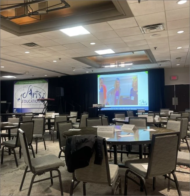 Empty conference room with round tables and chairs, a large screen displaying children on stage, and a banner for arts education in the background.