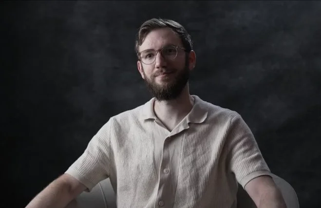 A man with glasses and a beard sitting against a dark background, wearing a light-colored short-sleeve shirt.