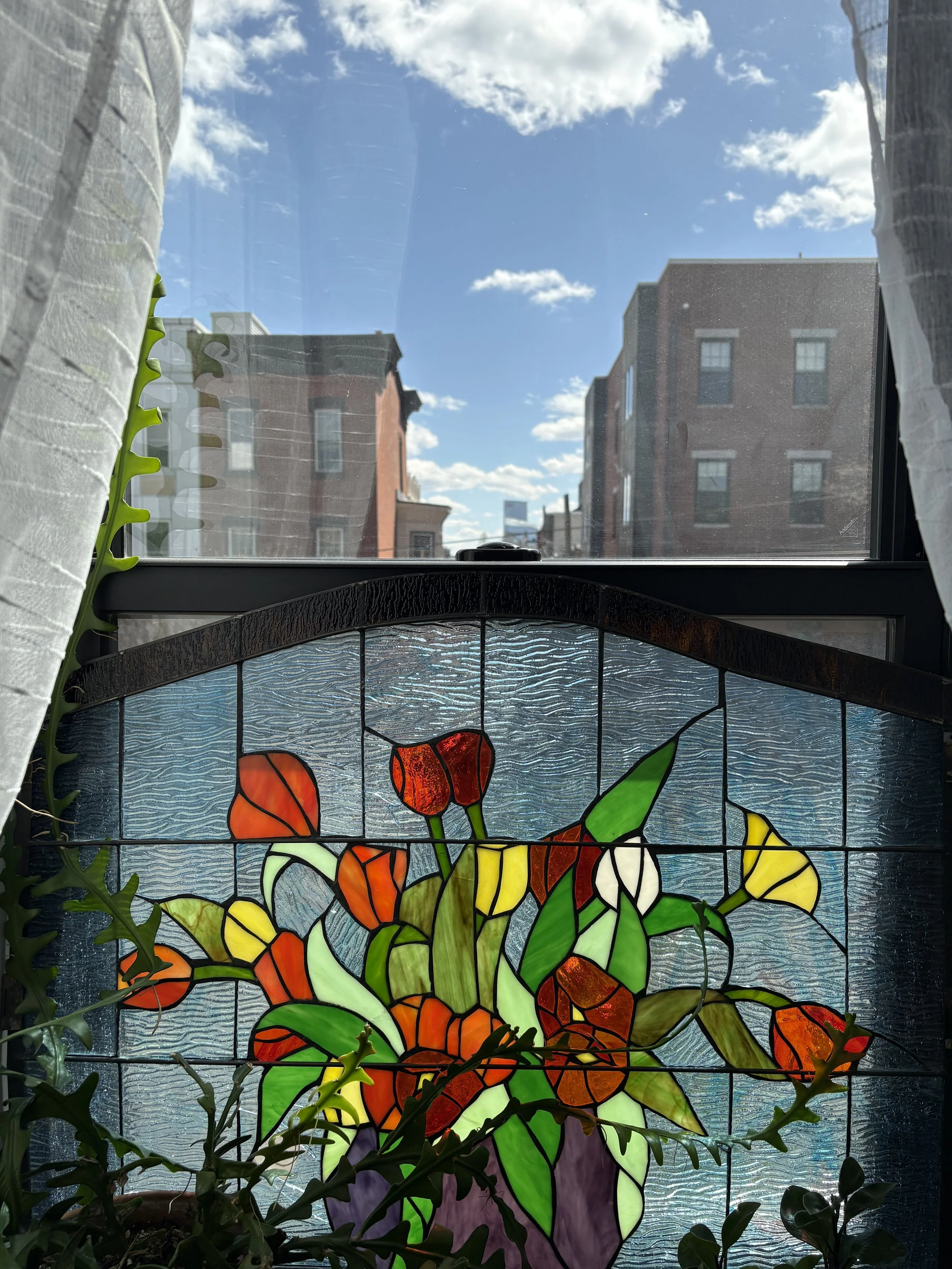 Colored stained glass flower arrangement with red, yellow, orange, and green flowers and leaves, indoors near a window with white curtains, sunlight shining through, city buildings in the background.