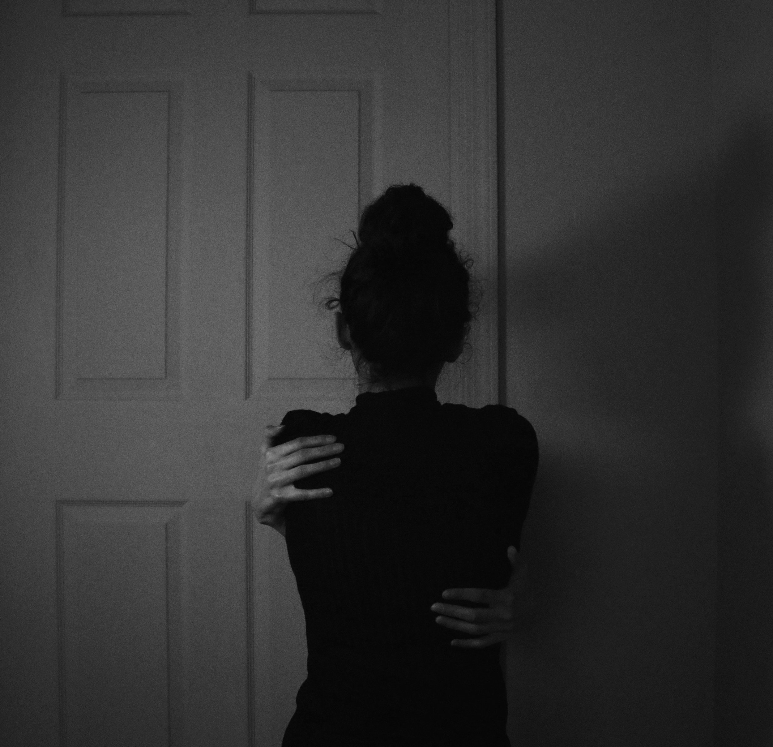 A woman facing a closed door, hugging herself with both arms, in a dimly lit room, image suggests symbolism of trauma and distress.