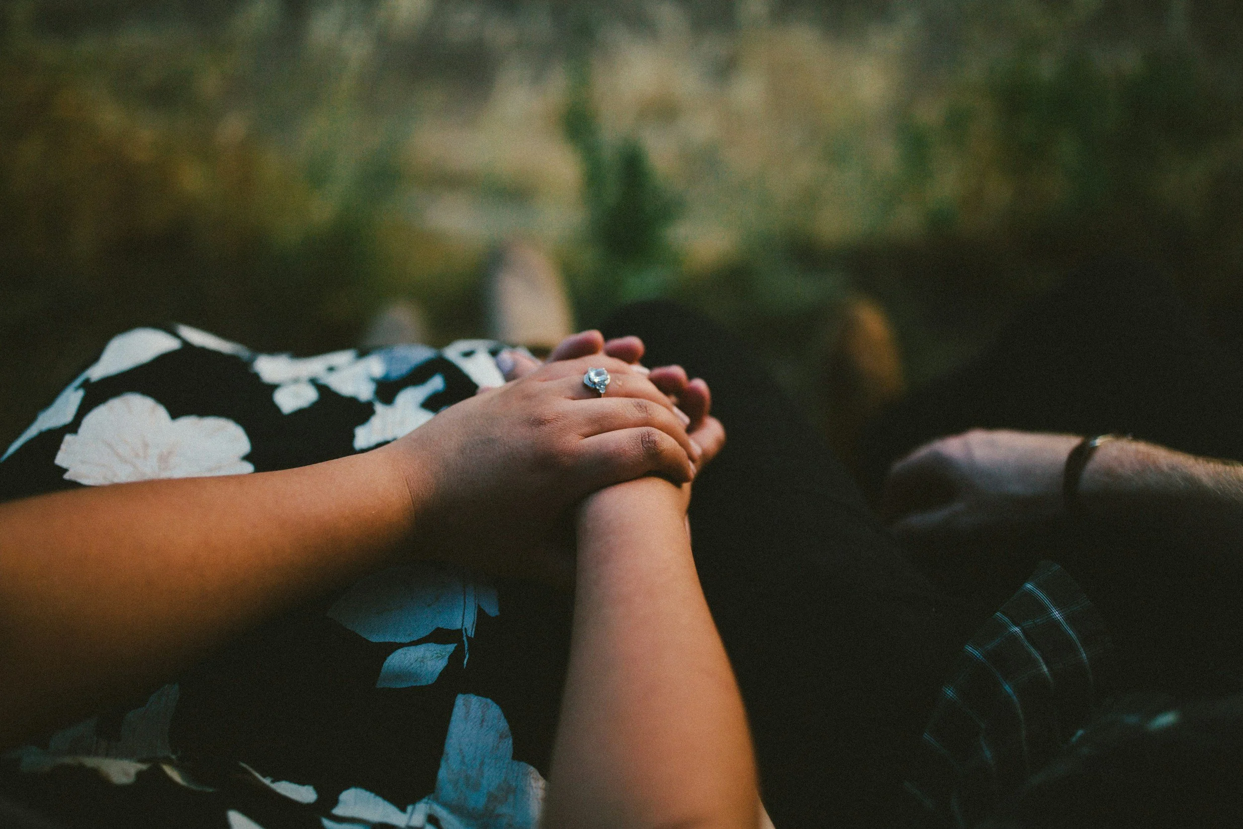 A person holding hands with another person, sitting outdoors with blurred grass and rocks in the background.