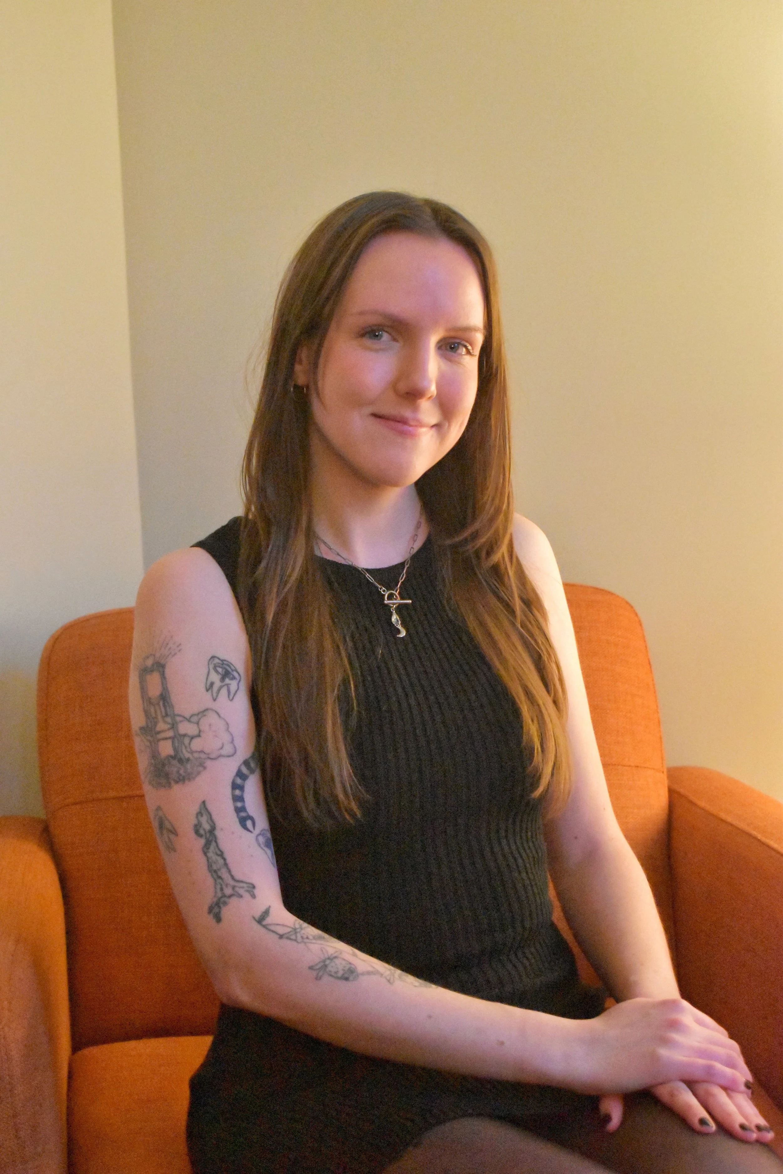 Female therapist in Philadelphia wearing a black top, sitting on an orange couch in a room with soft lighting and beige walls, smiling at the camera.