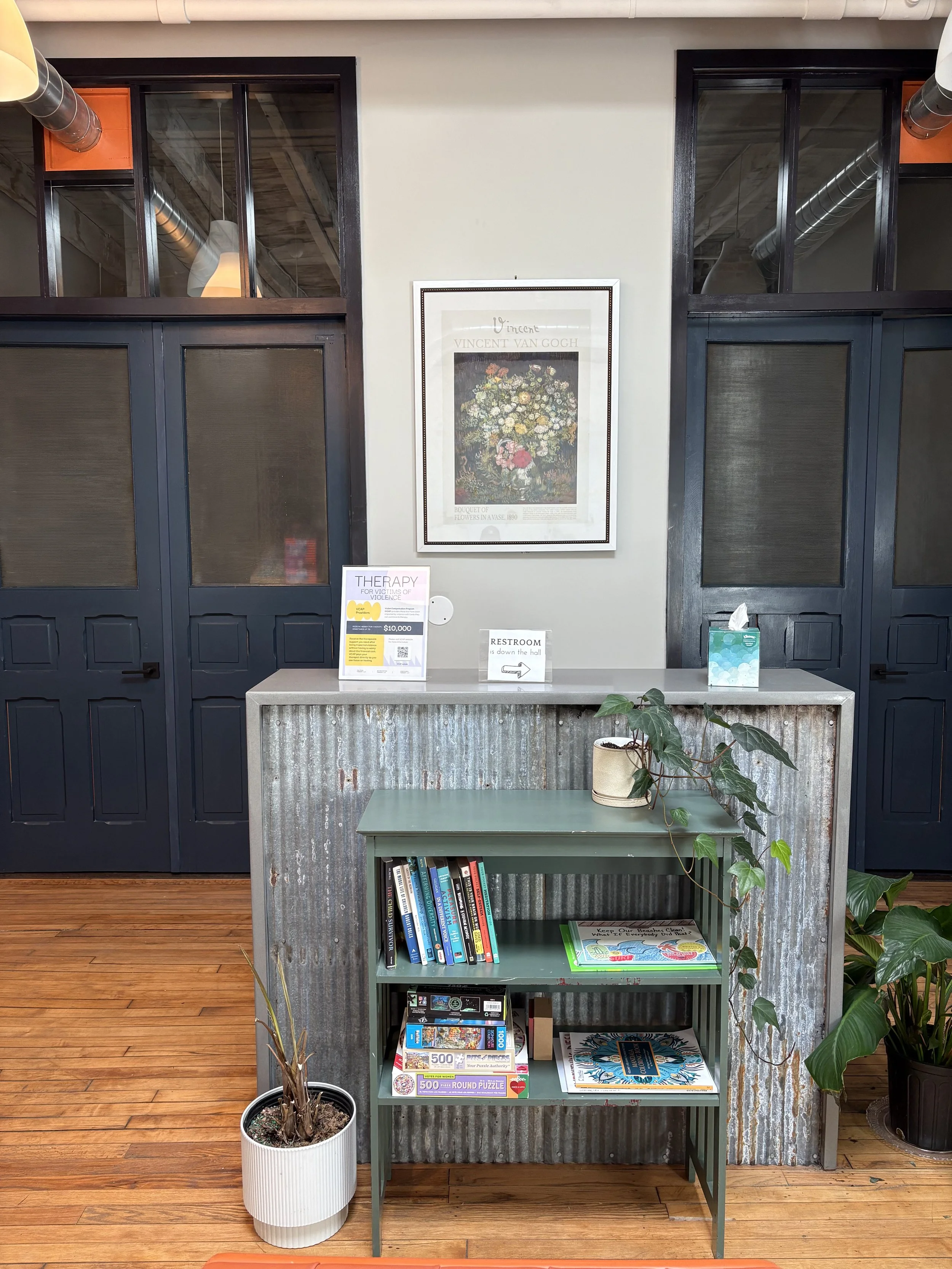 Indoor reception area with a framed Vincent van Gogh flower poster on the wall, a corrugated metal counter with therapy and restroom signs, a small green bookshelf with books and puzzles, and potted plants on a wooden floor.