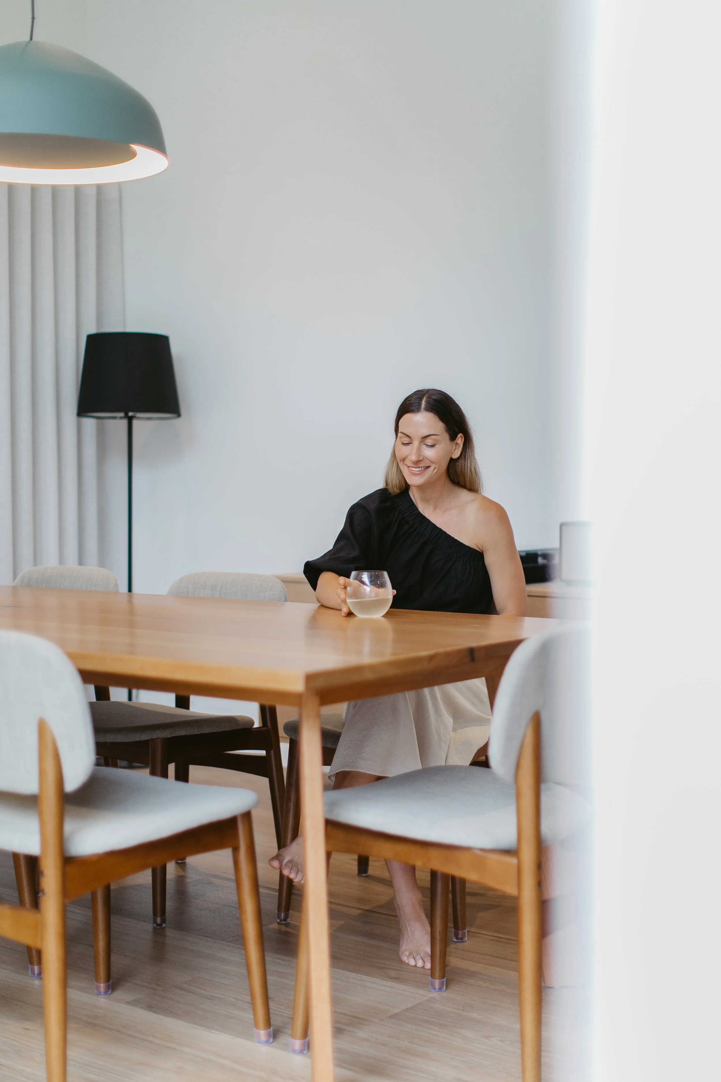 A woman sitting at a dining table, holding a glass of water, smiling, with bare feet, in a bright, modern room with wooden floors and minimalist decor.
