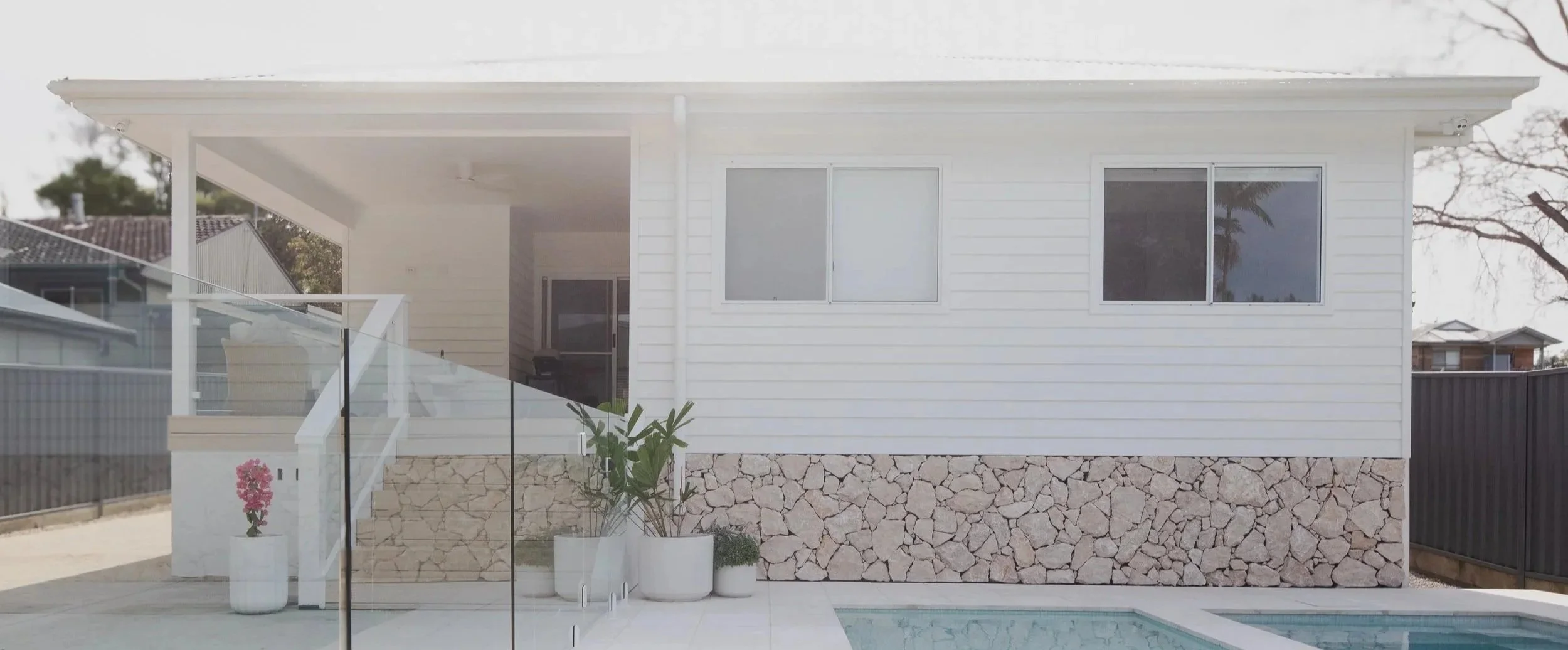 Modern white house with a stone base, two windows, a small swimming pool, potted plants, and a staircase with glass railing leading to a porch.