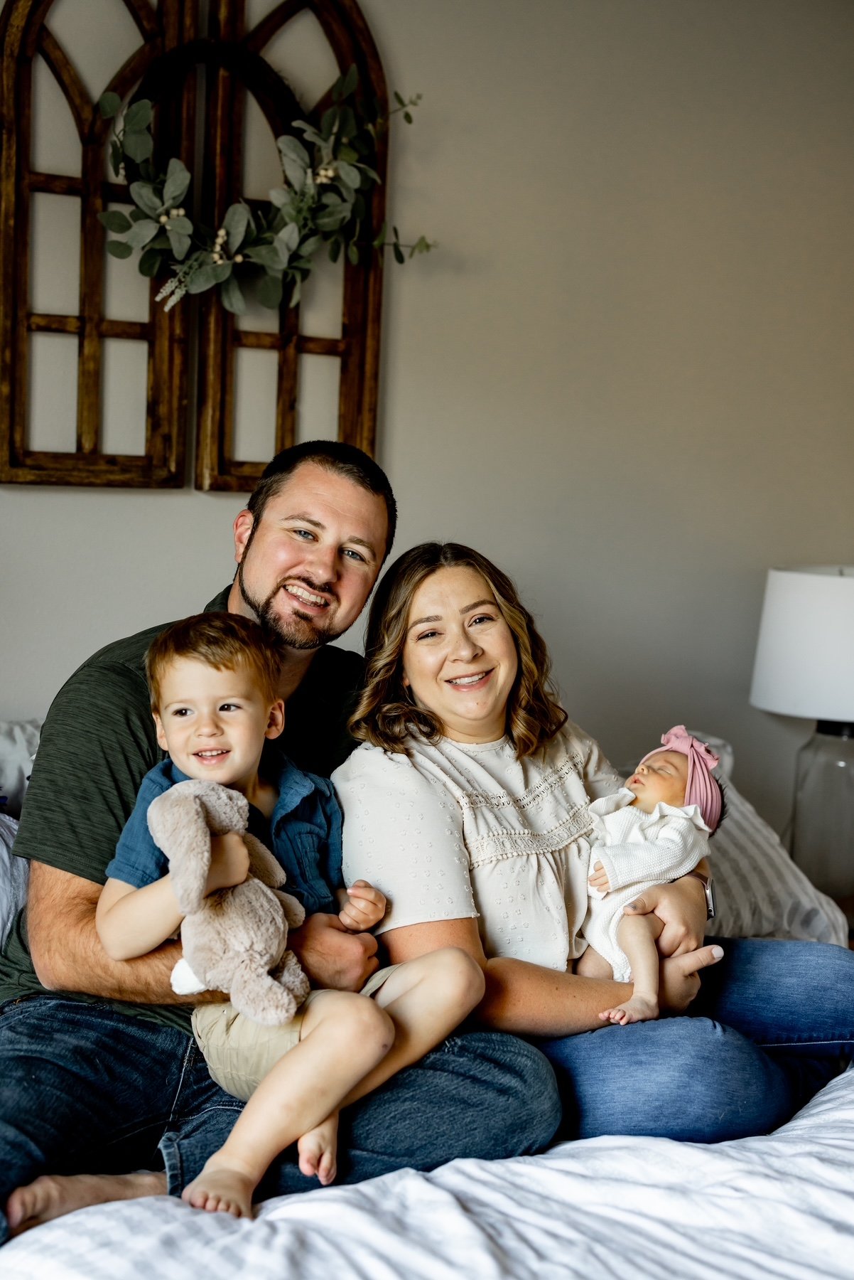 A family of four sitting on a bed, holding their two young children, smiling at the camera. The father has a beard and dark hair, the mother has medium-length brown hair and a light-colored blouse, the boy has red hair and is holding a stuffed animal, and the baby girl is wearing a pink headband with a bow.
