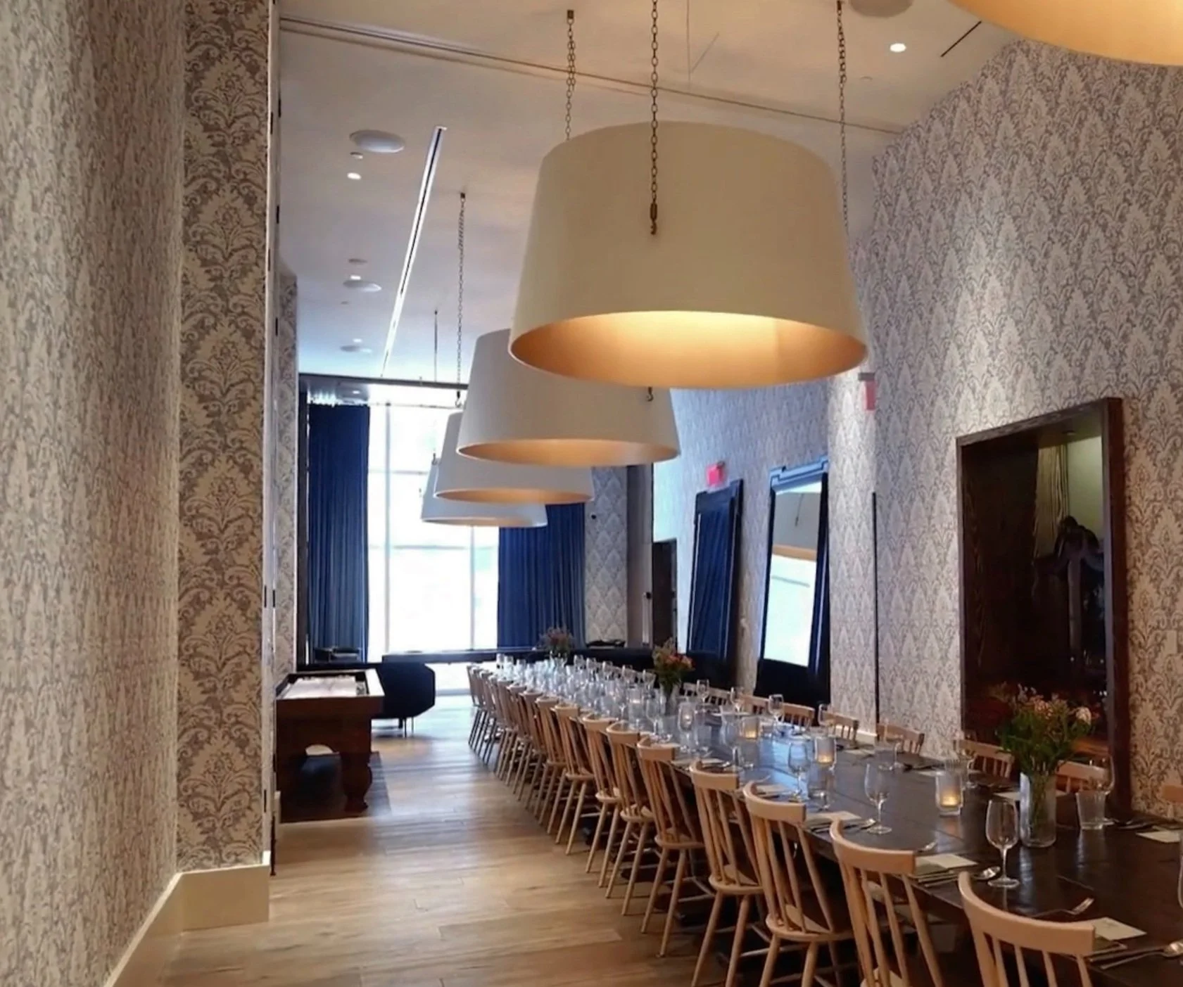 Elegant dining room with long table, wooden chairs, large pendant lights, decorative wallpaper, and natural lighting from windows.
