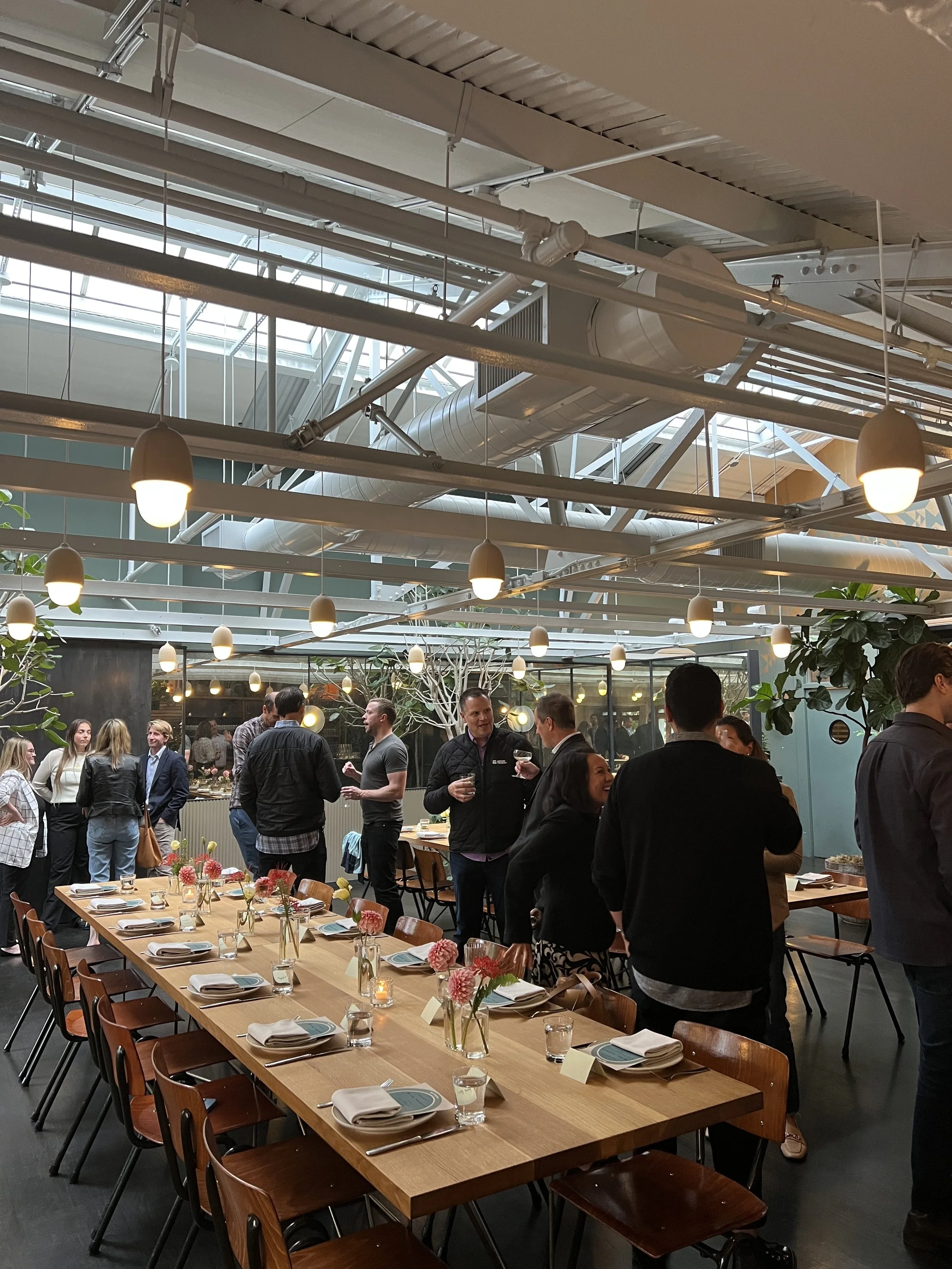 Large dining room with set tables and people socializing, overhead industrial-style lighting, and potted plants.