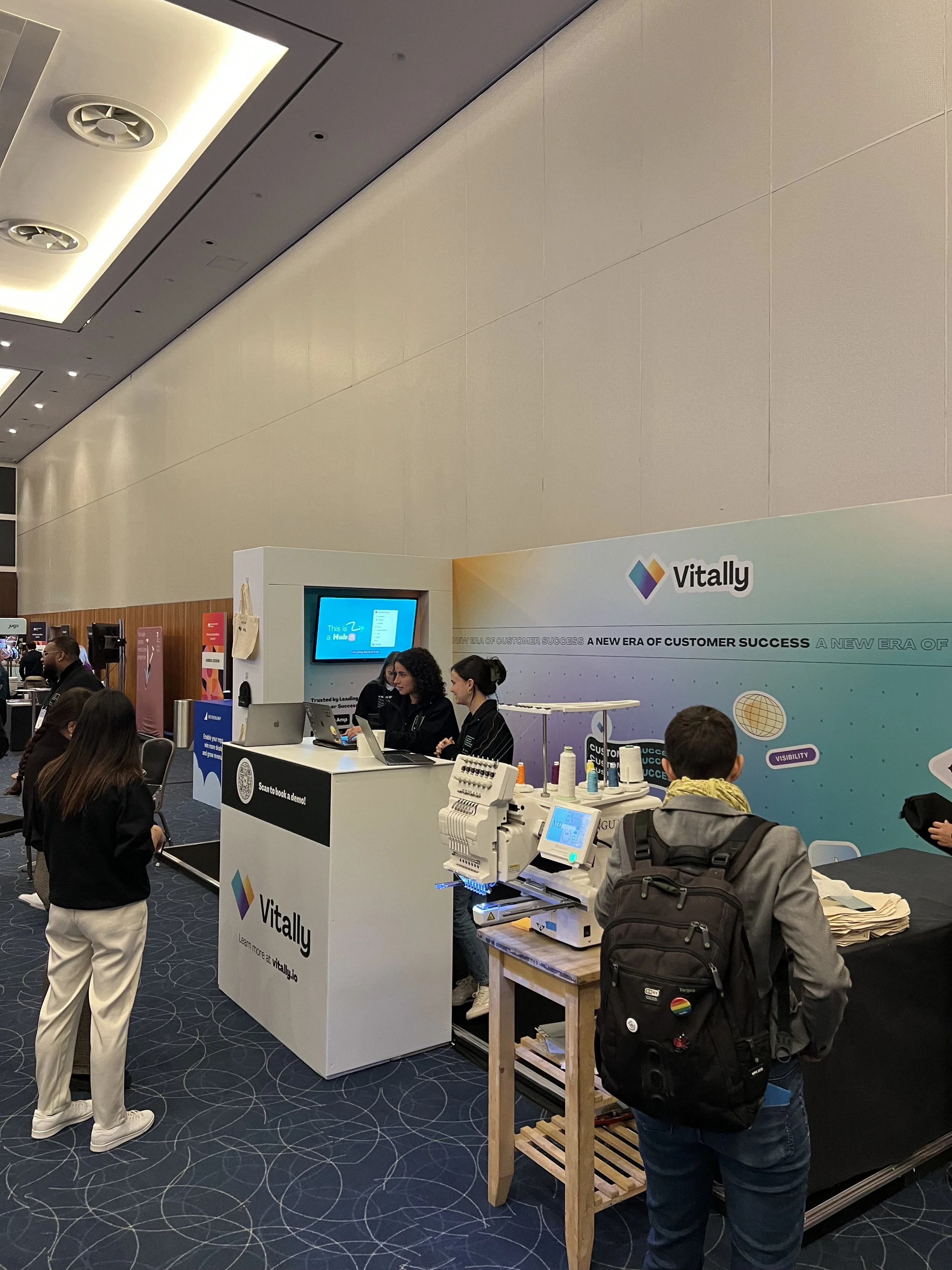Exhibition booth with Vitally branding, staff interacting with visitors, embroidery machine displayed, and promotional materials setup in a large conference hall.