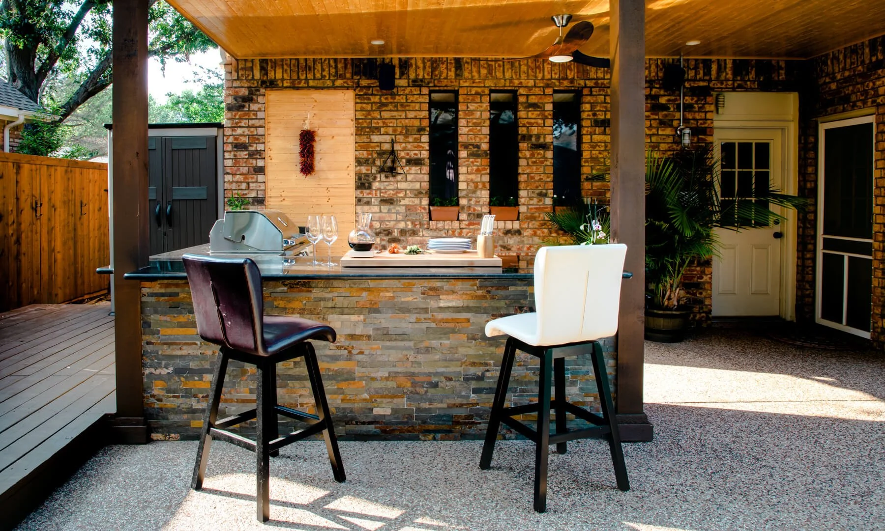 Remodeled backyard patio in Urbandale, Iowa showcasing a curved paver patio with built‑in grill, sink and countertop, a circular stone seating wall, and a round wicker dining set with green cushions and colorful place settings.