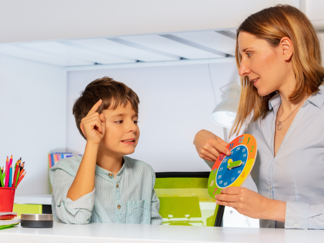A young boy is sitting at a desk with an adult woman during a tutoring session. The woman holds a colorful teaching clock, pointing to it while explaining something to the boy inside a bright room.