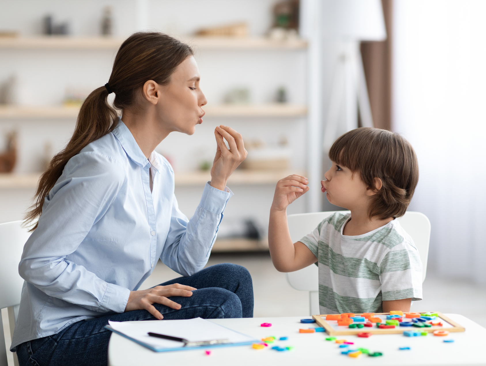 A woman and a young boy playing a game together at a table, both making gestures with their hands and blowing bubbles towards each other, in a bright room with shelves in the background.