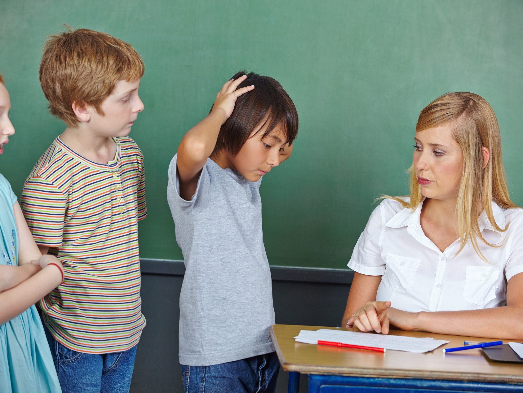 A classroom scene with a teacher and three students. The teacher, a woman with blonde hair, is sitting at a desk with papers and pens. One student, a boy in a gray shirt, is touching his head as if in distress. Two other students, a girl in a light blue dress and a boy in a striped shirt, are standing next to him, looking concerned.