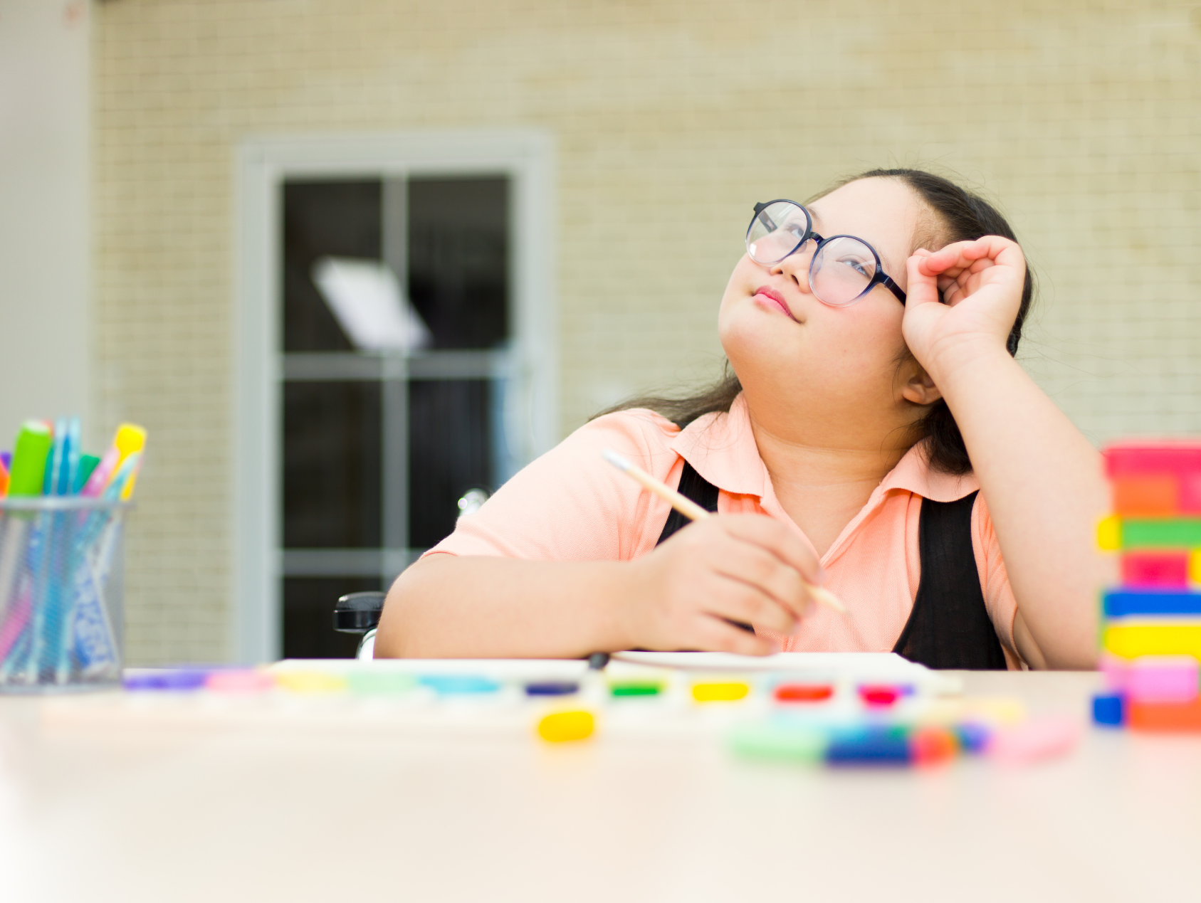A young girl with glasses, wearing a peach-colored shirt and black suspenders, sitting at a table with colorful markers and paper, looking thoughtfully upward with her hand resting on her head.