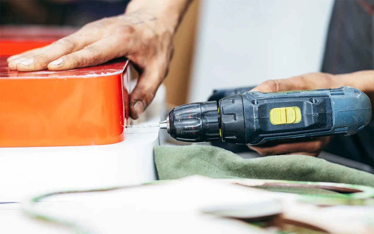 Person using a cordless drill to fasten a red object on a white surface, with a green cloth nearby.