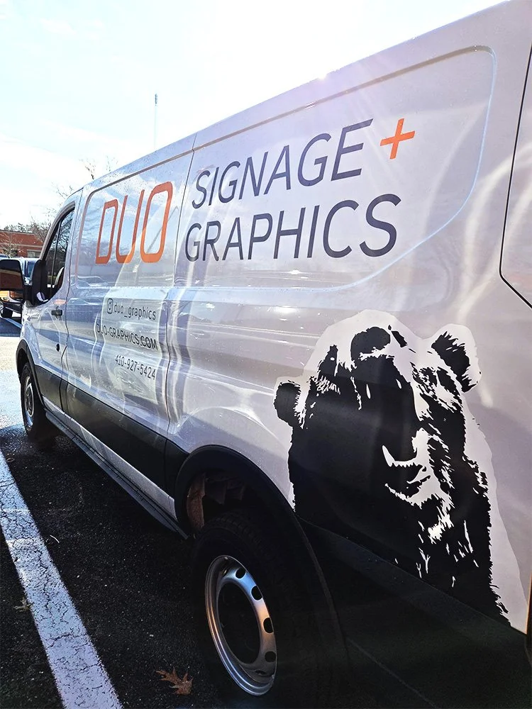 Side of a white van with signage and graphics for Duo Signage and Graphics, including company details and a black lion head graphic.