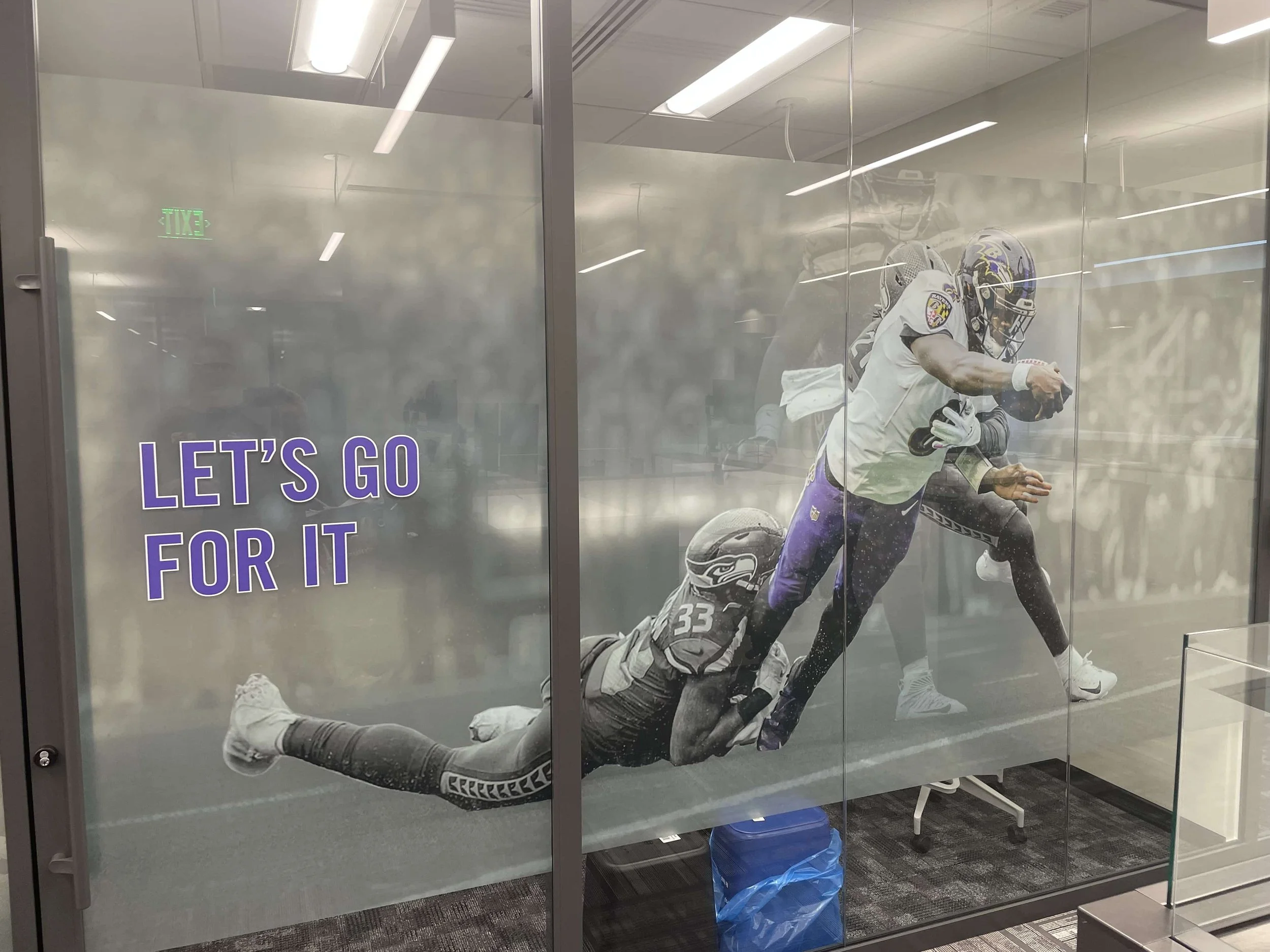 Glass wall with a sports poster showing football players. One player is diving with the ball, while another is reaching for it. The poster has a purple text that says, "LET'S GO FOR IT."