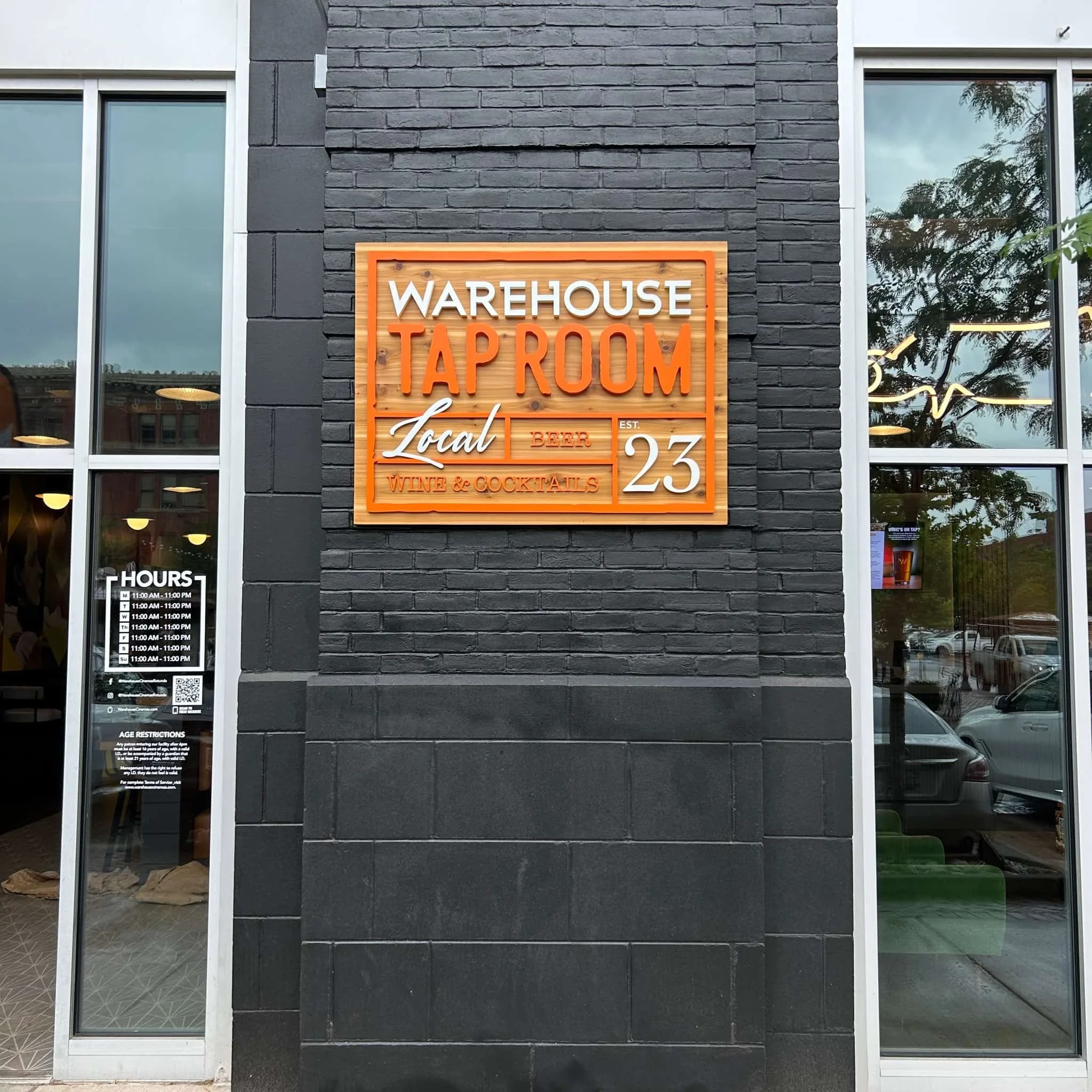 Exterior of a building with a sign that reads 'Warehouse Tap Room, Local Beer, Wine & Cocktails, Est. 23' on a black brick wall, with large glass windows showing inside and outside parking lot.