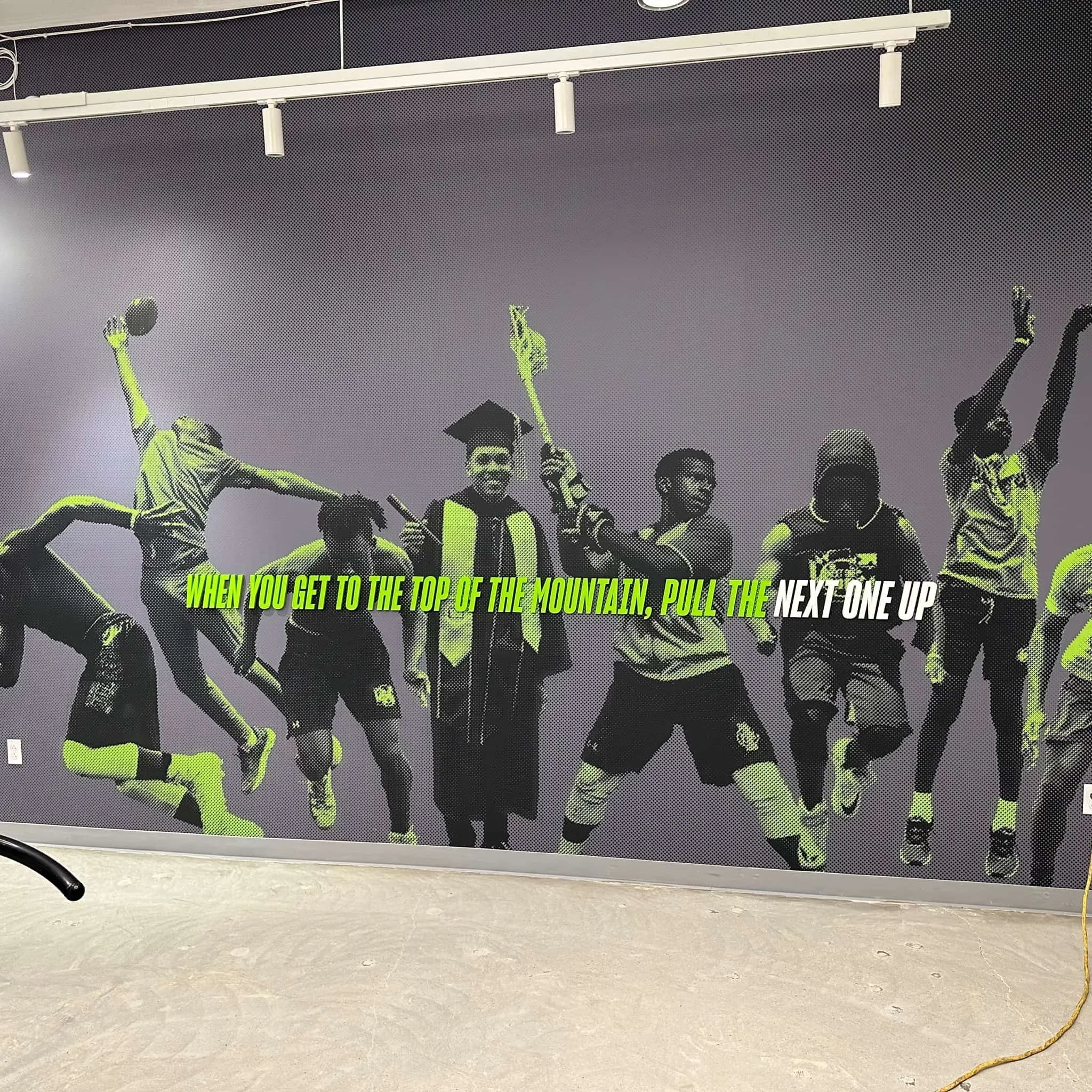 Wall mural of diverse young people celebrating, with one wearing a graduation cap, and a motivational quote in bold green and white text, reading: 'When you get to the top of the mountain, pull the next one up.'
