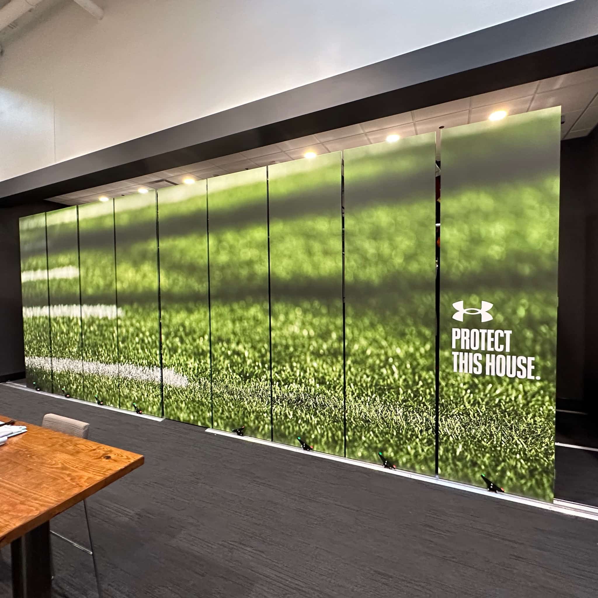 A large, multi-panel display wall with an image of green grass and the Under Armour logo along with the text "Protect This House." in a room with dark walls and a wooden table in the foreground.