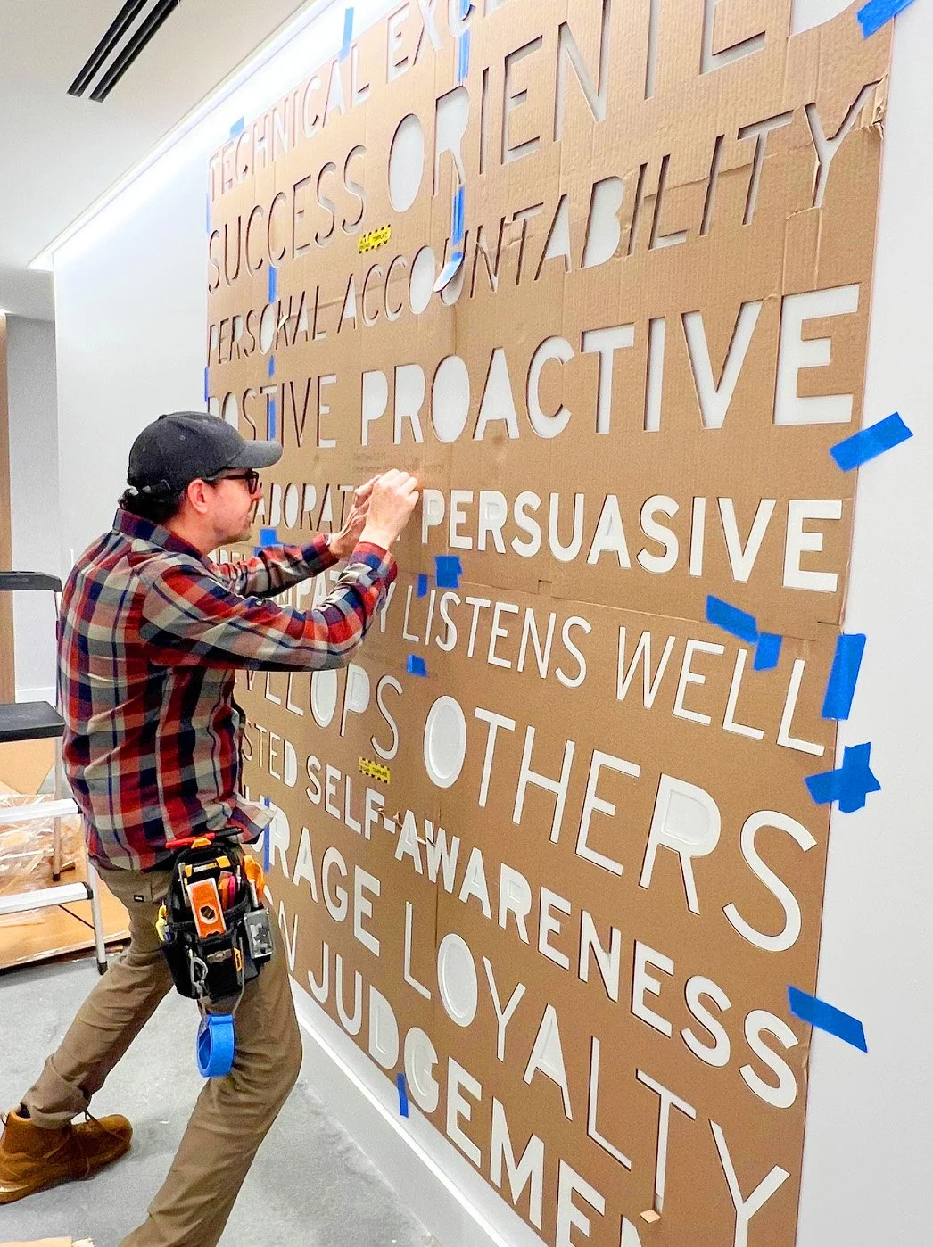 A person working on a large collaborative wall with phrases cut out from cardboard, taping and arranging them. The background is a plain white wall.