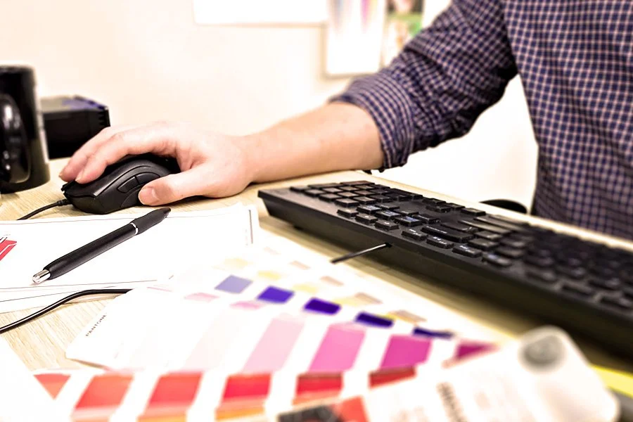 Person using a computer mouse at a desk with color swatches, a pen, and a keyboard.