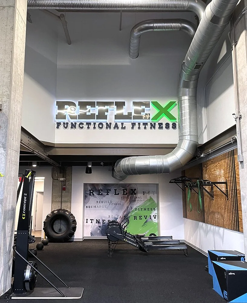 Interior of a fitness gym with a illuminated sign reading REVLEX FUNCTIONAL FITNESS, and fitness equipment including a tire, rowing machines, and pull-up bars.