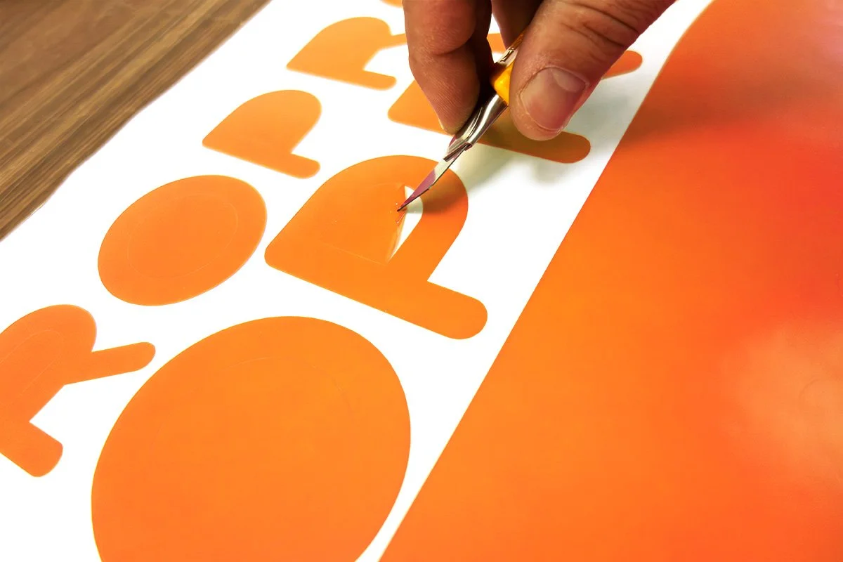 A person using a craft knife to cut out orange letters from a paper sheet.