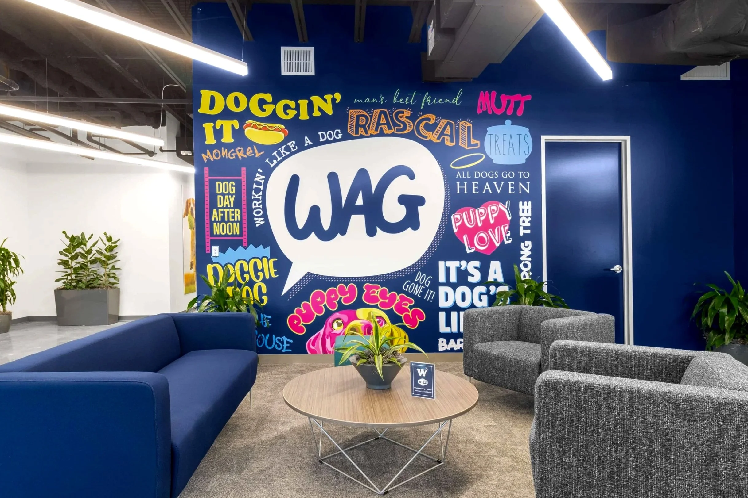 Colorful wall mural with dog-themed words and phrases such as "Wag," "Doggin' it," "Rascal," "Puppy Eyes," "Puppy Love," and "All dogs go to heaven." The mural decorates a modern office lounge with a blue wall, gray and blue couches, a round wooden table, and a potted plant.