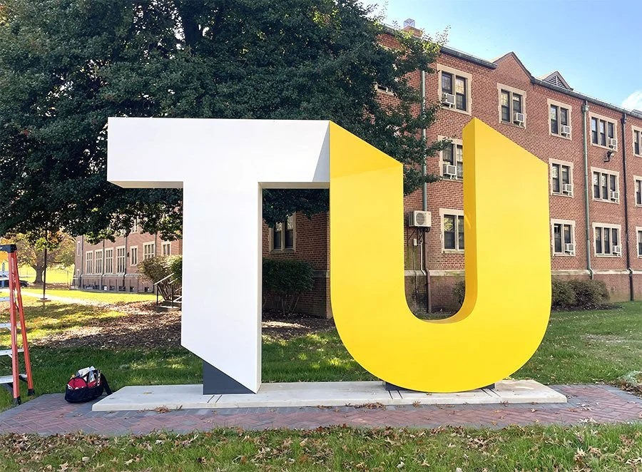 Large 3D letters spelling out 'TU' in white and yellow, placed outdoors on a concrete base near a brick building with multiple windows, trees, and grass nearby.