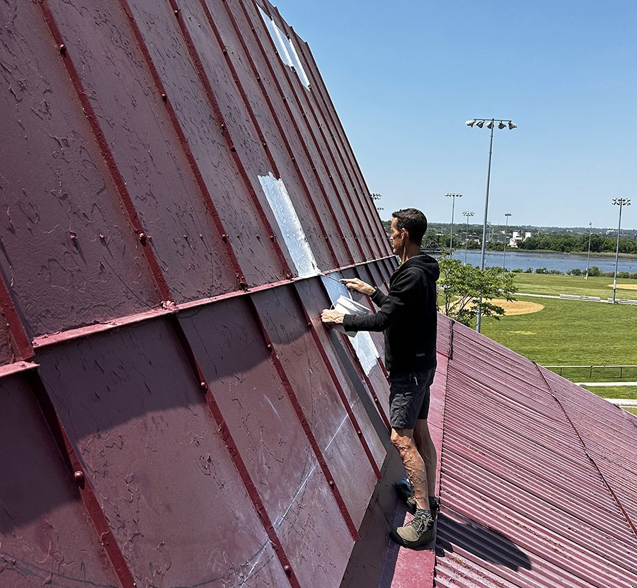A man in black clothing on a steep red metal roof painting or repairing the surface on a sunny day.