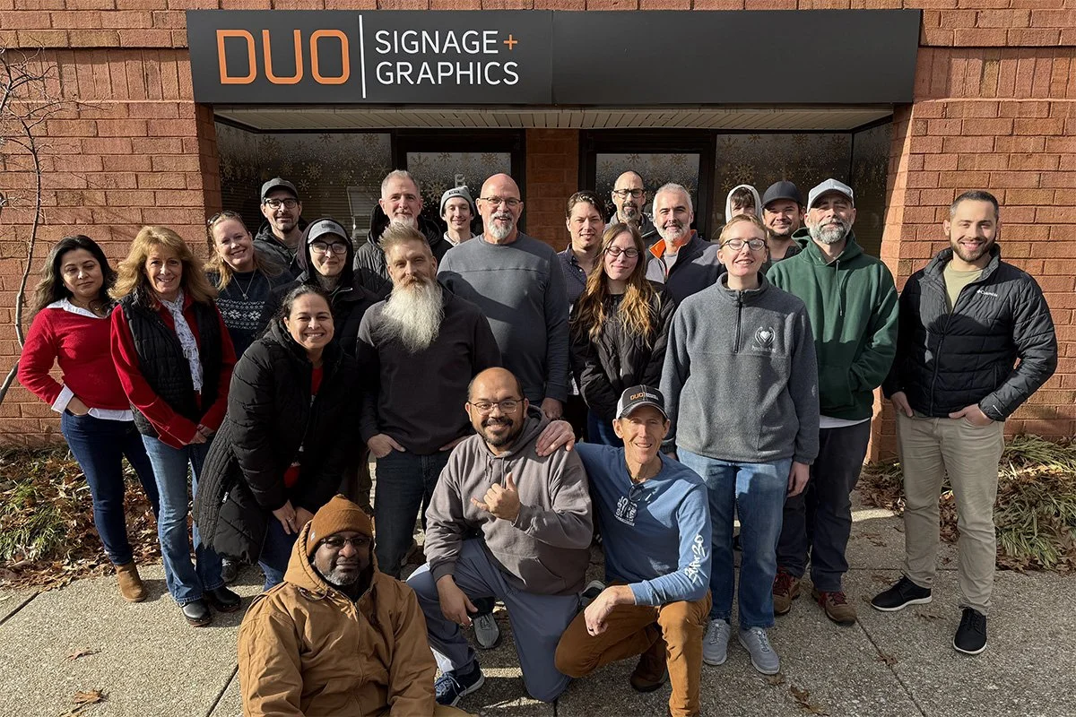 Group of people standing outside a brick building with a sign that reads 'DUO Signage + Graphics'