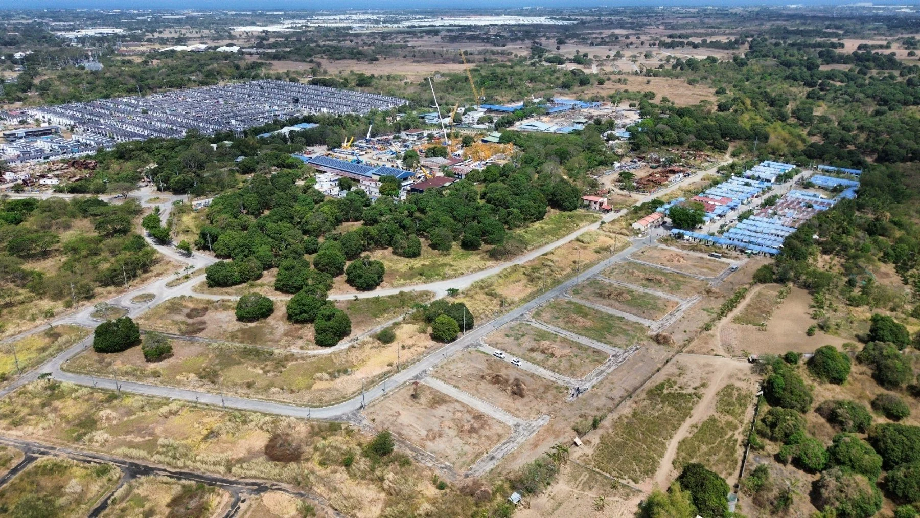Aerial view of a construction site with surrounding greenery, roads, and residential structures. There are cranes, buildings under development, and partially developed plots. The area is bordered by dense vegetation and open fields.