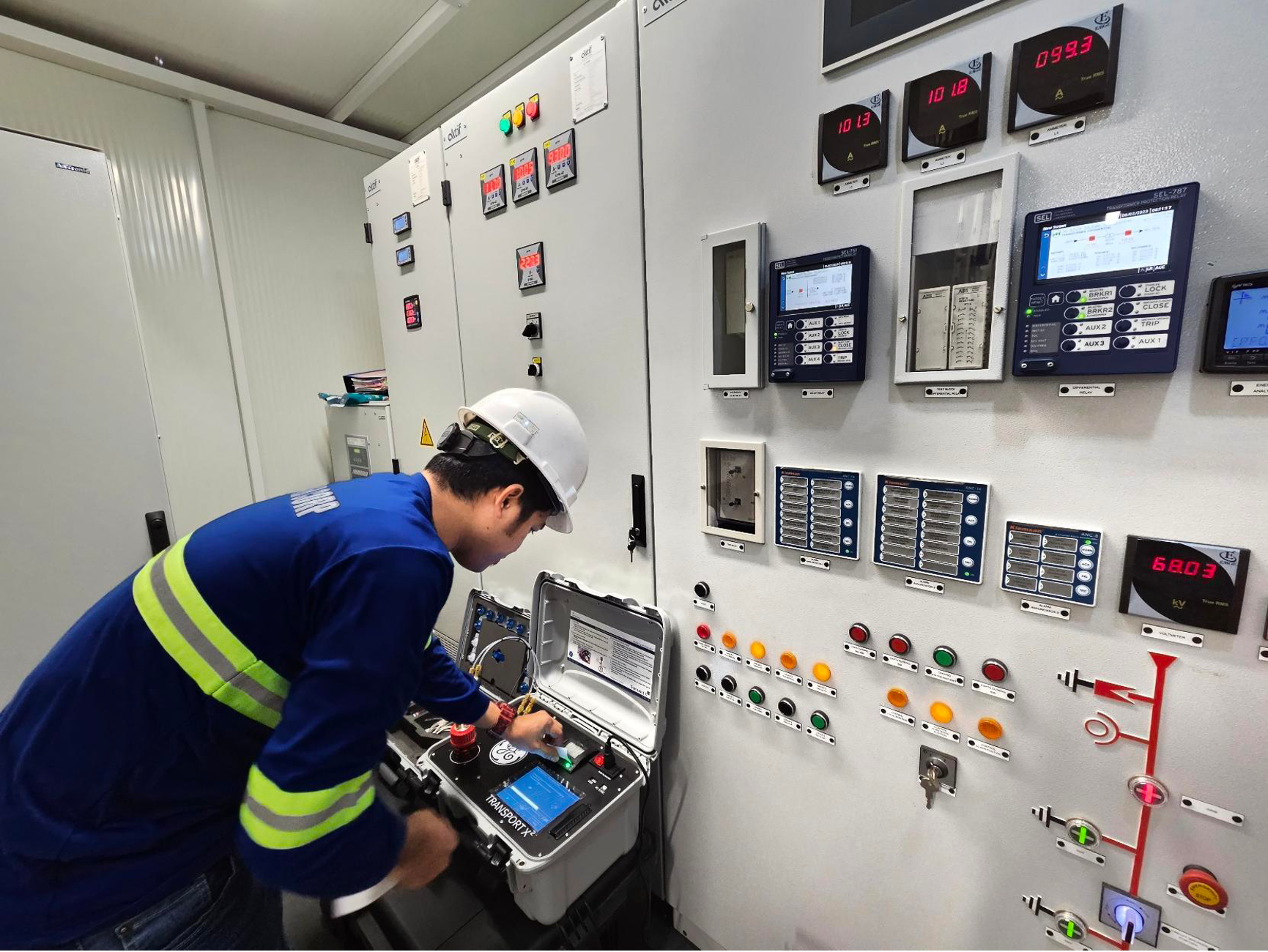 Technician operating an industrial control panel with various digital displays, buttons, and switches, wearing a hard hat and safety gear.