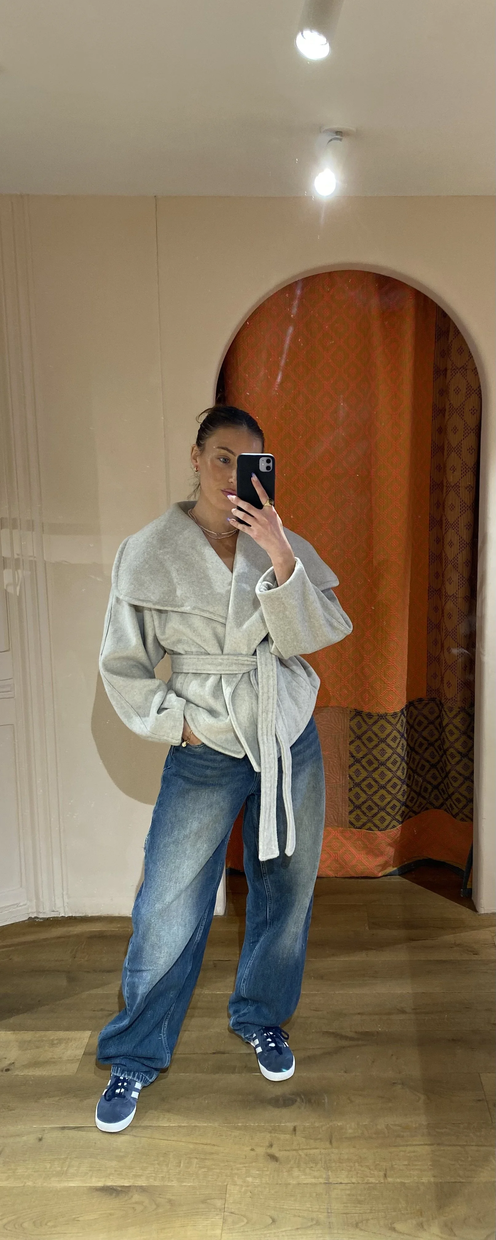Woman taking a mirror selfie, wearing a light gray belted coat, loose blue jeans, and navy sneakers in a room with wooden floors and orange patterned curtains.