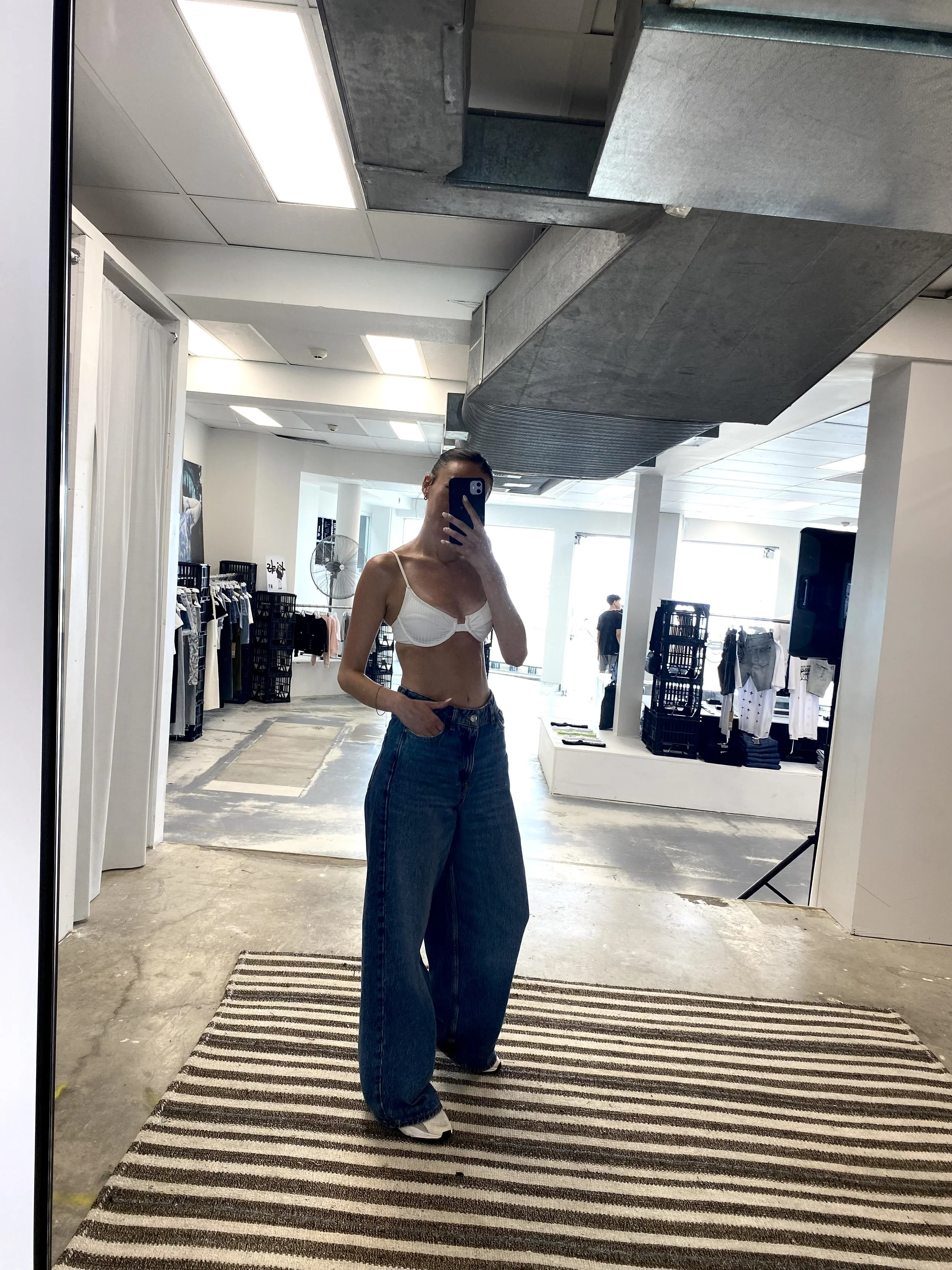 Woman taking a mirror selfie in a clothing store, wearing a white lingerie top and large blue jeans.