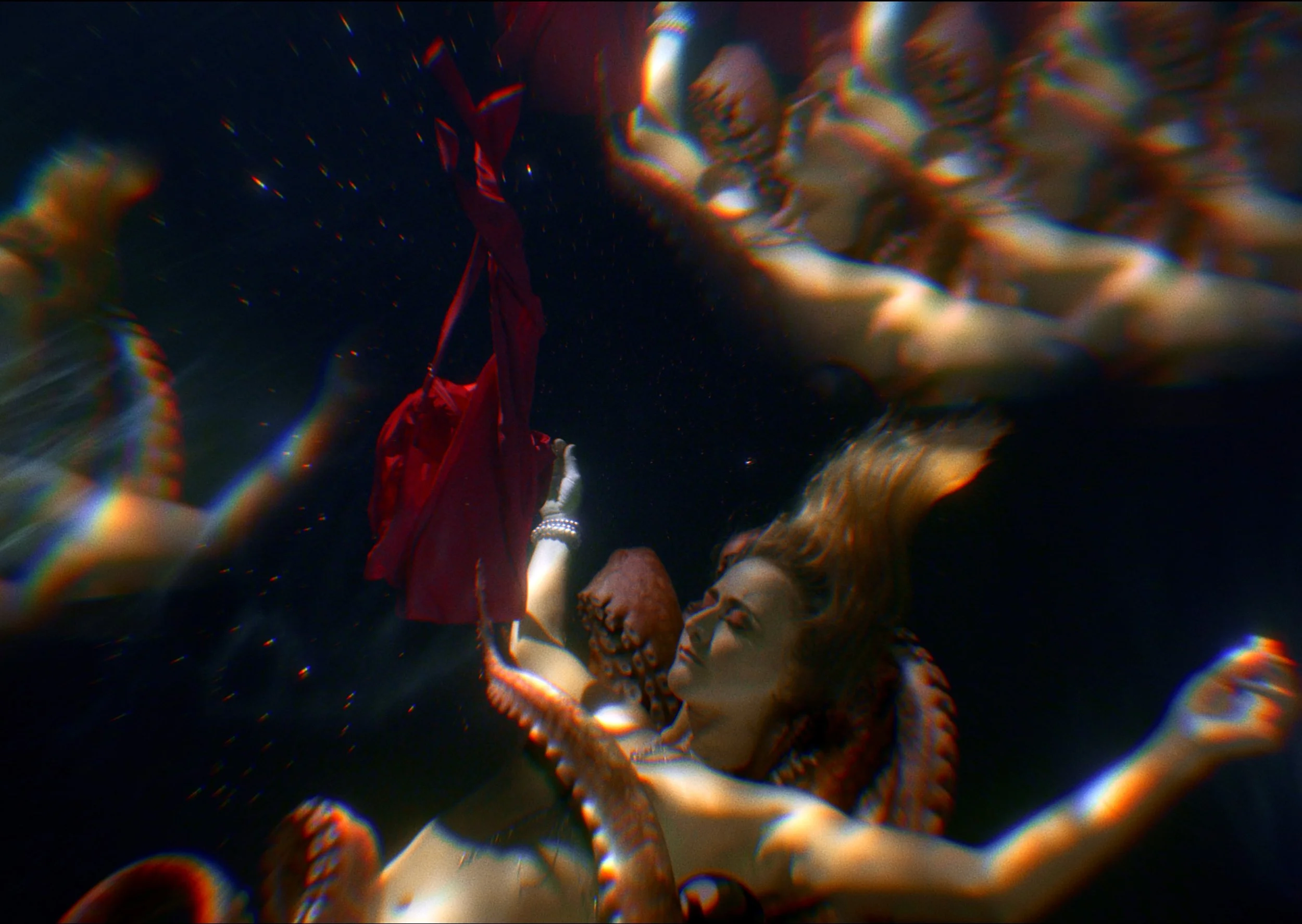 A woman with long, brunette hair underwater, holding a red piece of clothing in her right hand, surrounded by octopus tentacles.