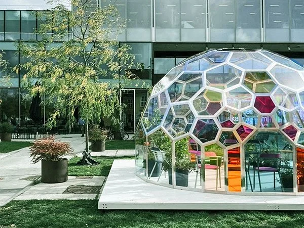 En Njord kuppel og uderum med klare og farvede glas på en træterrasse foran en moderne bygning, omgivet af træer og planter