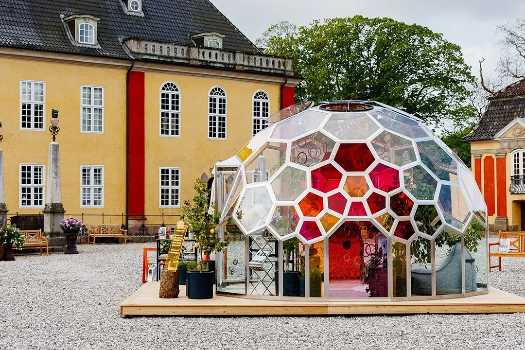 En Njord kuppel og uderum med klare og farvede glas, på en stenbelagt plads foran historiske bygninger, indeholdende planter og indretning.