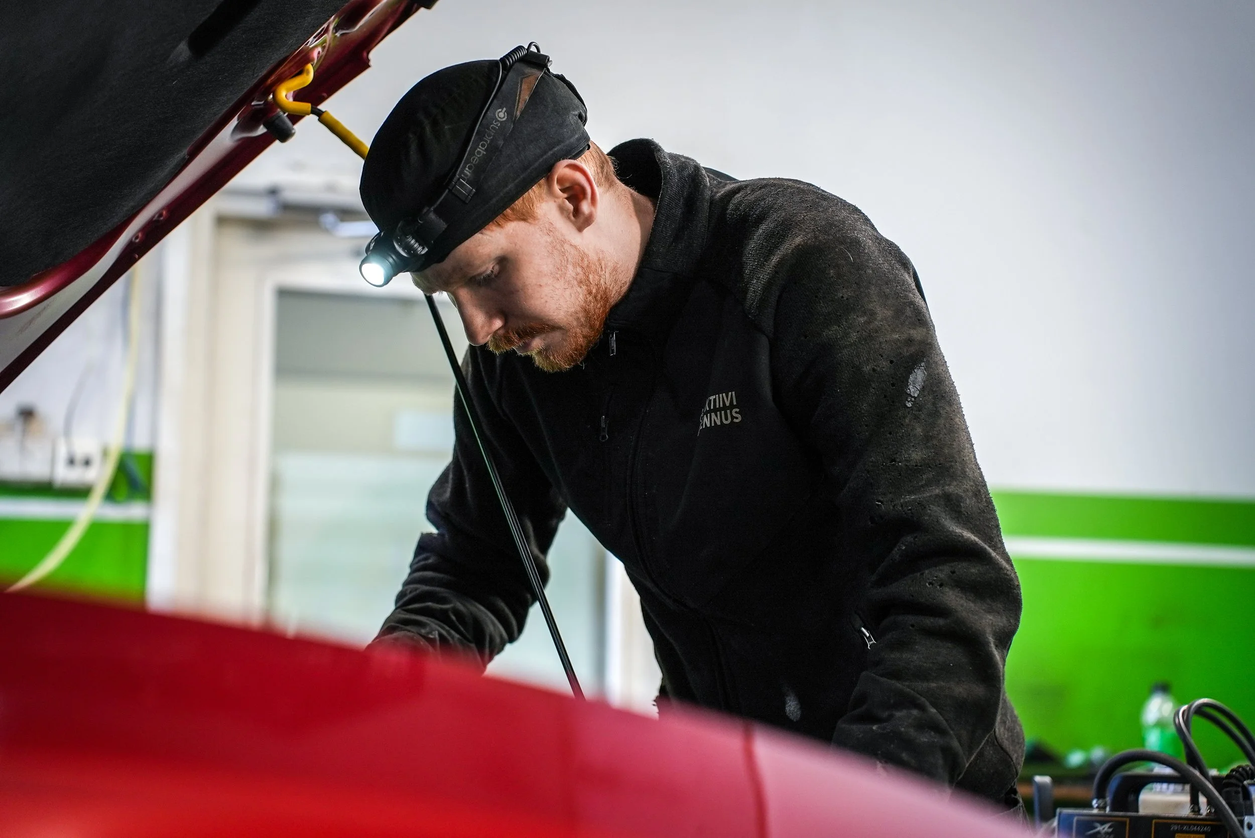 Mies työskentelee autokorjaamossa, katsoo läheltä auton moottoriosaa ja hänellä on otsalamppu päässä.
