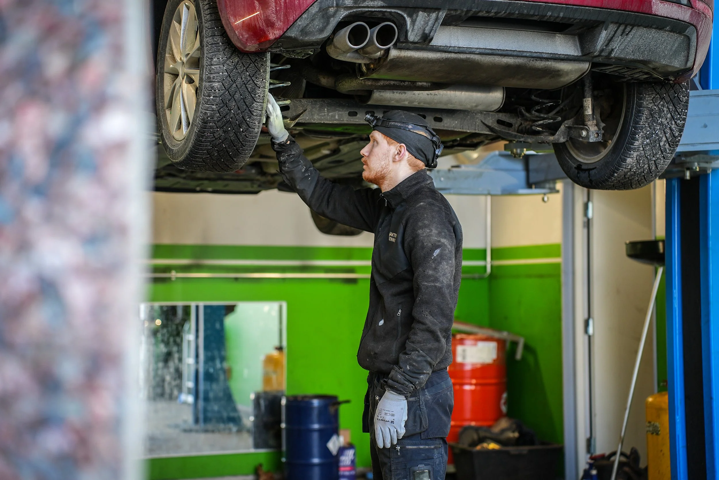 Automekaanikko työskentelee nostetun auton alla korjaamolla, tarkkailee ja korjaa auton osia.