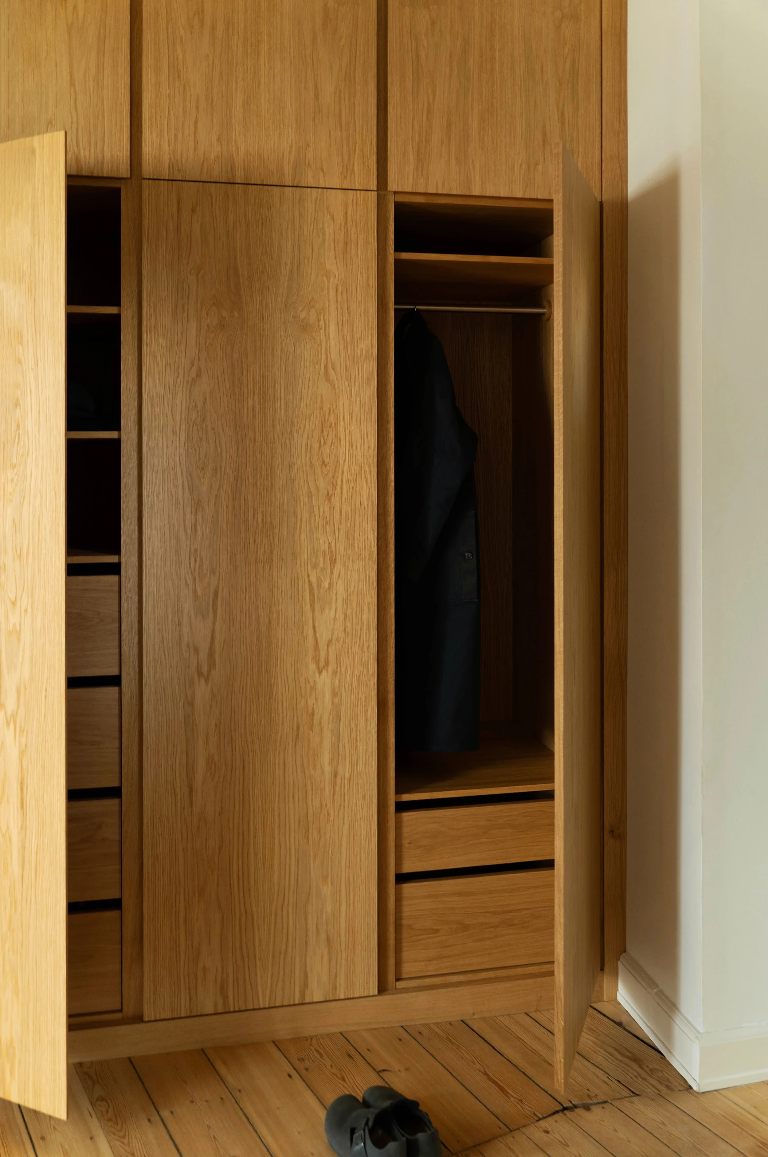 An open wooden wardrobe with a single dark coat hanging inside, upper shelves above, and three drawers at the bottom. A pair of shoes is on the wooden floor in front of it.