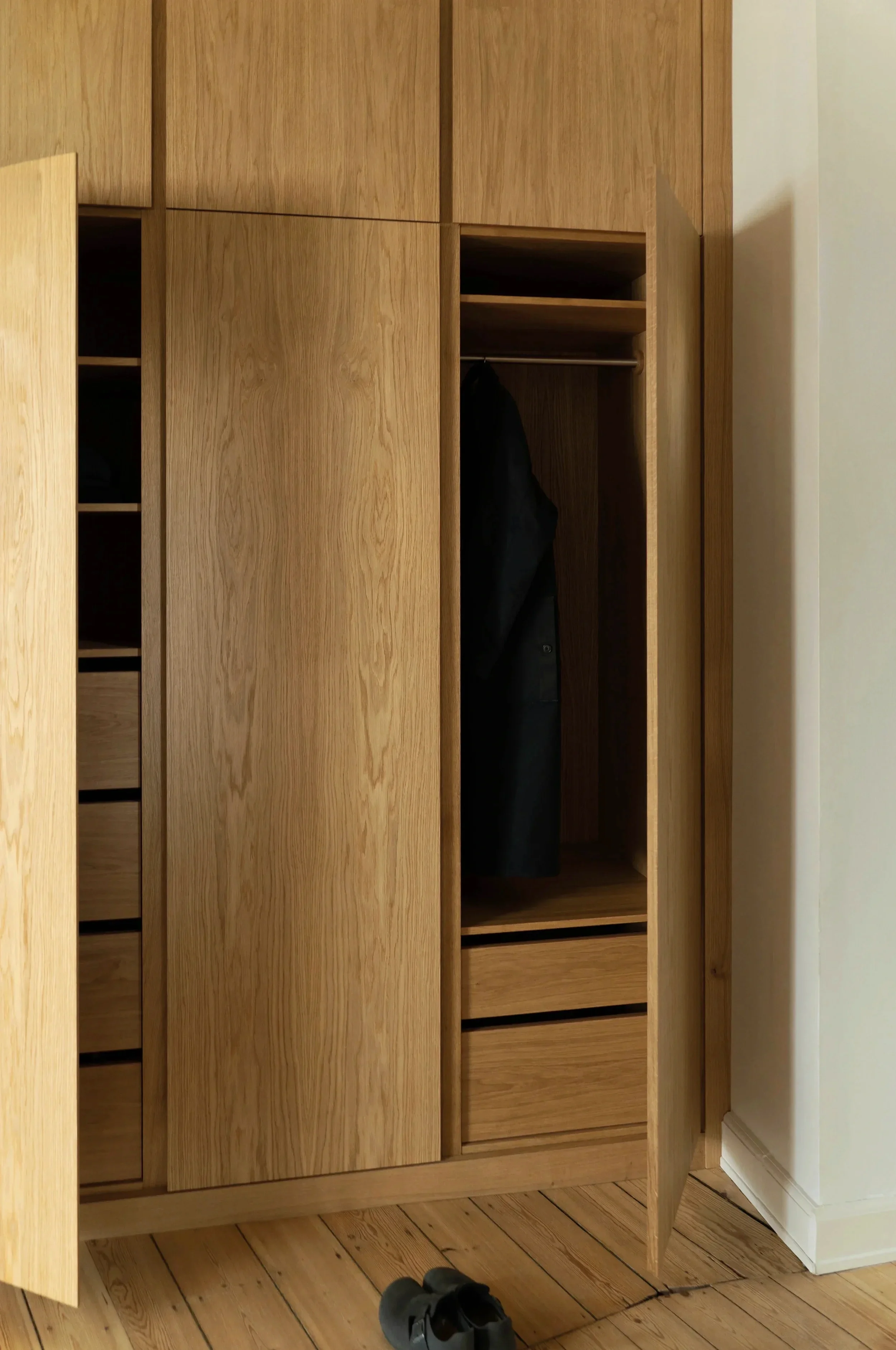 Open wooden wardrobe with a black coat inside, standing on a wooden floor with black shoes in front.