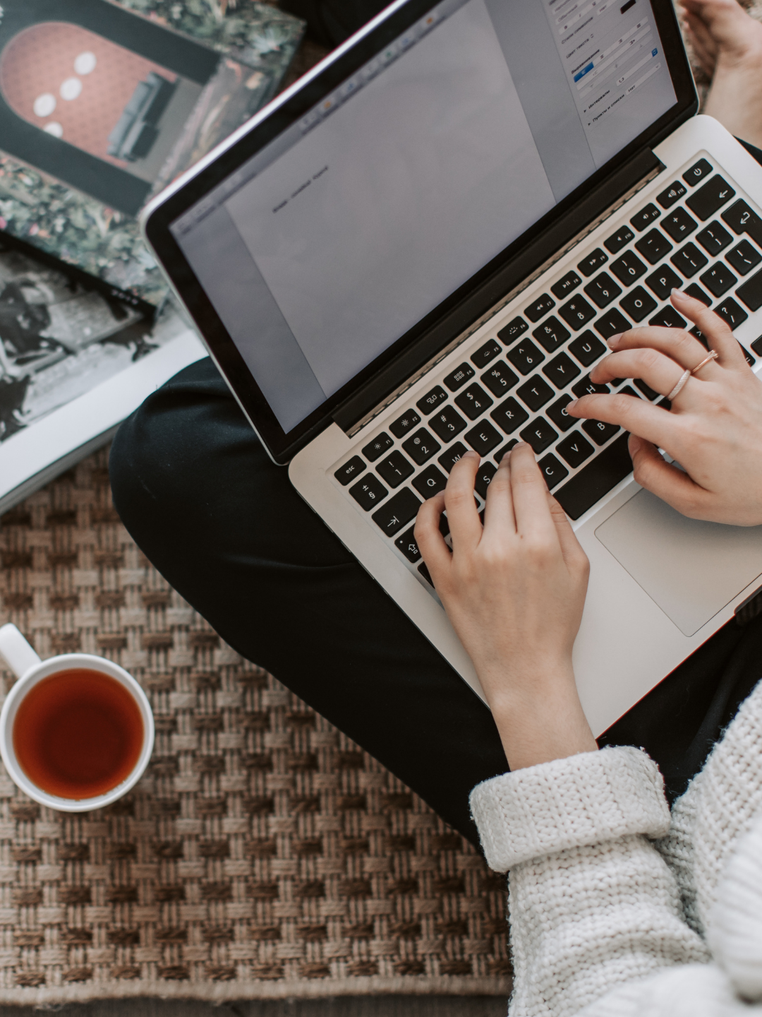 Person using a silver laptop with a black keyboard, sitting on a woven mat, with a cup of tea nearby and photo books in the background. Image used to describe step three in the onboarding process for Socials With Michelle which includes my work