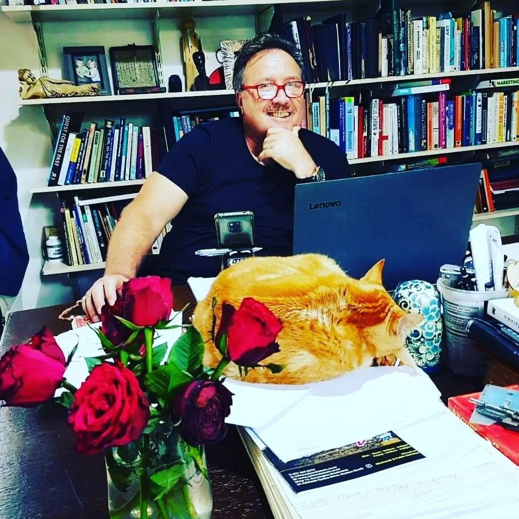A man with glasses and a beard sitting at a desk with a laptop, smiling and resting his chin on his hand. A sleeping orange cat is curled up on the desk among papers, a vase of red roses, and various office items. Behind him is a bookshelf filled with books and decorative items.