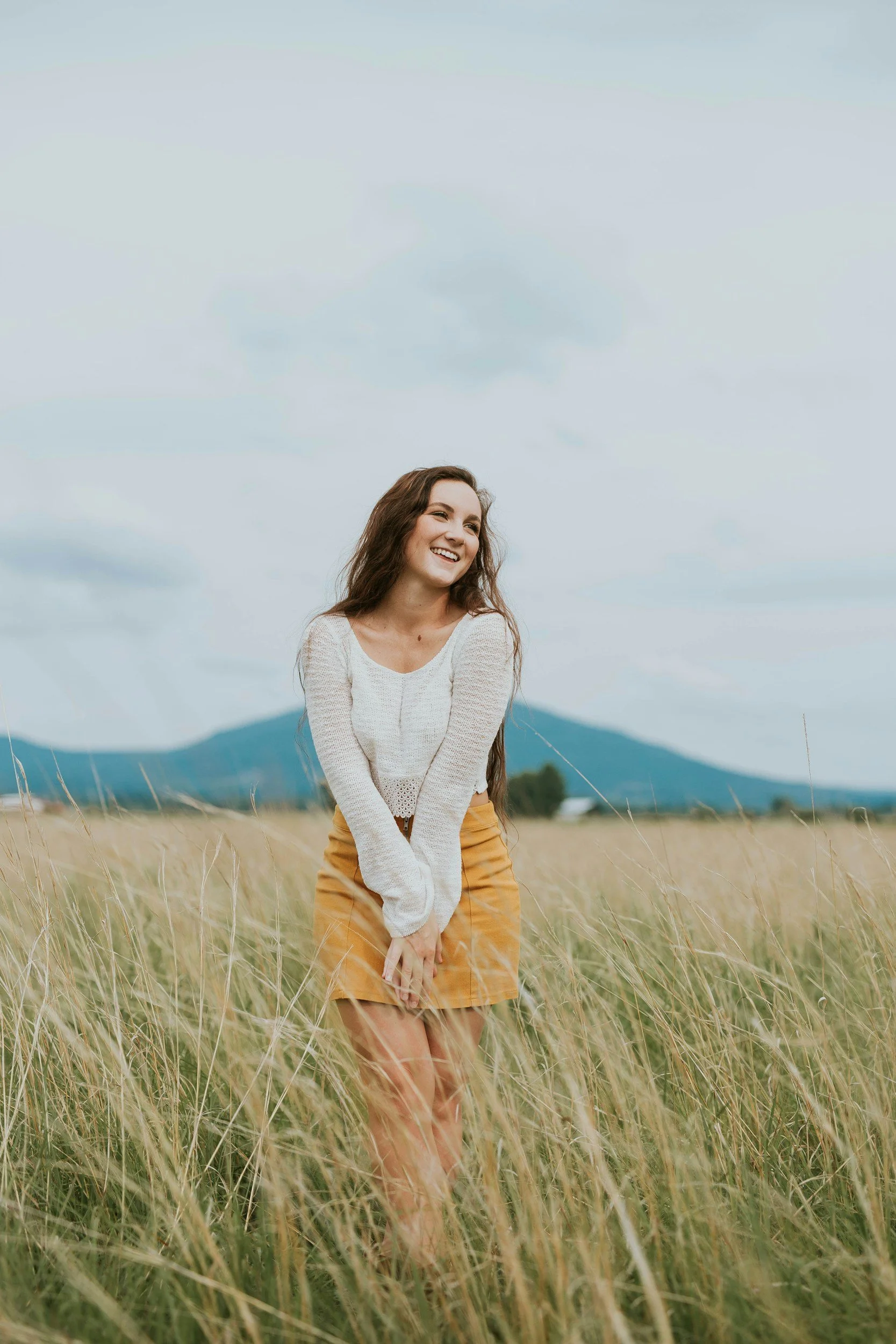 Woman in white sweater and yellow skirt smiling in a field of tall grass with mountains in the background.