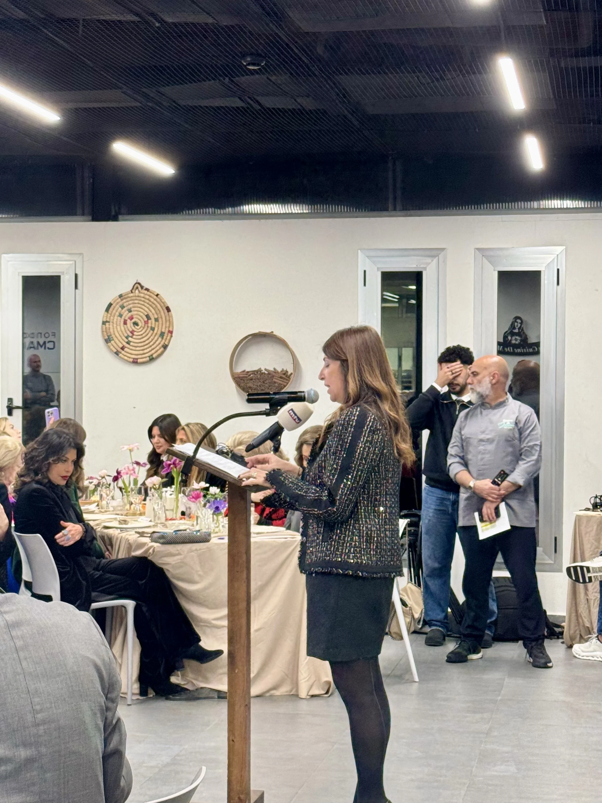 People socializing at a gathering, some holding wine glasses, in a room with stone walls and wooden shutters.