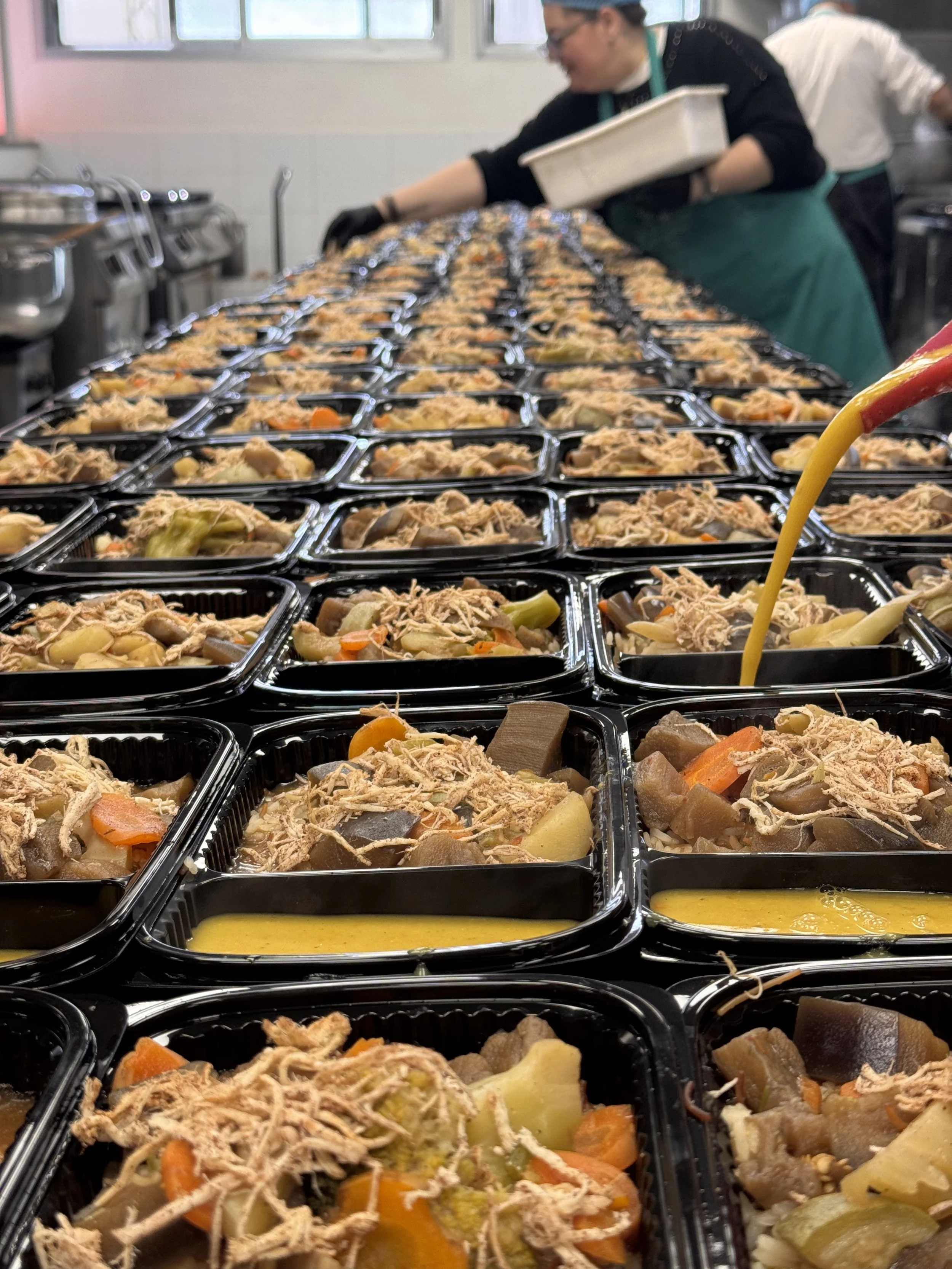 People working in a food preparation and distribution setting, wearing hairnets and gloves, packing food into black containers at a service station.